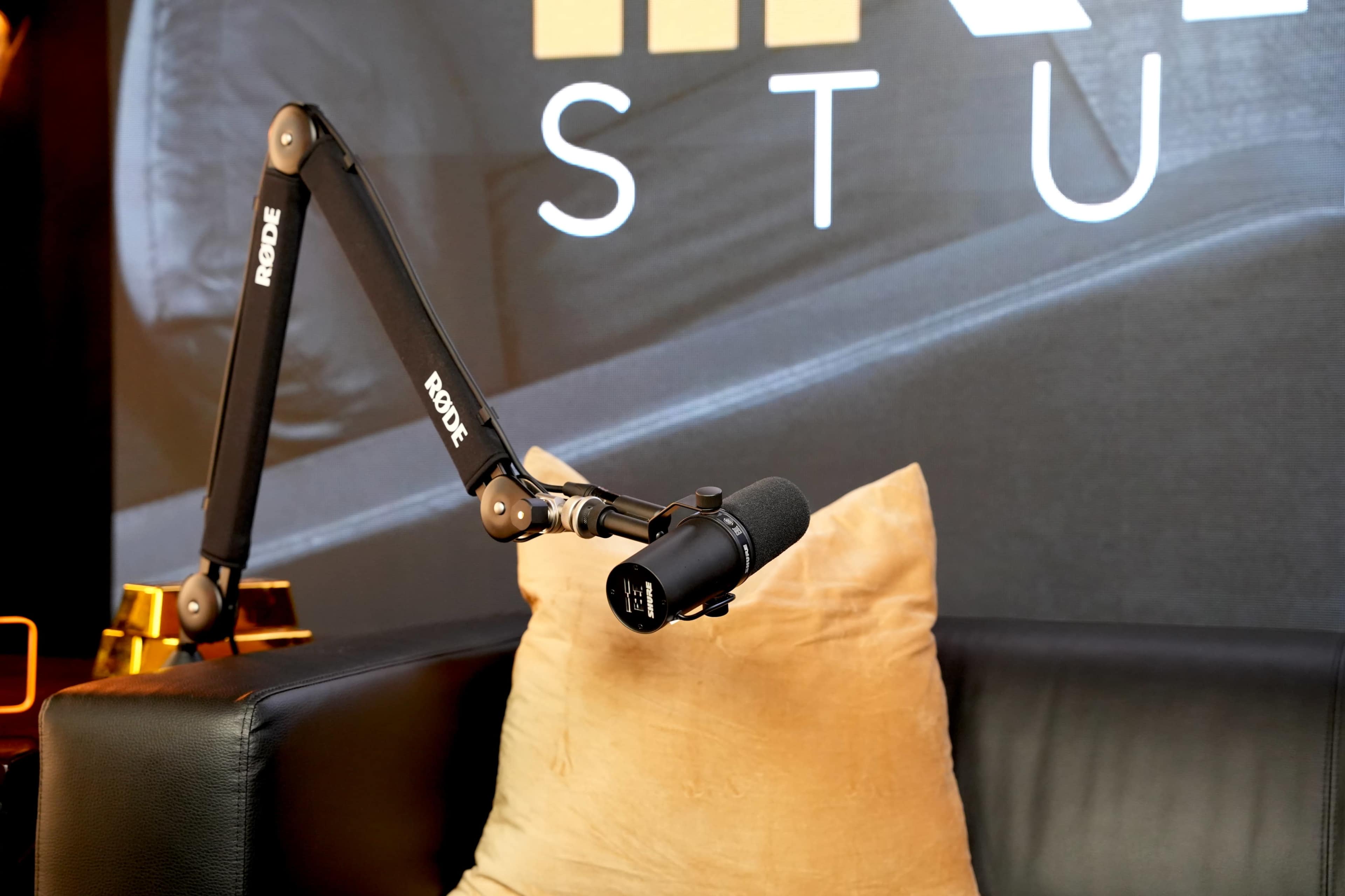 A Rode microphone is mounted on an adjustable arm on a black leather sofa with a tan pillow in front of a backdrop featuring "STUDIO."