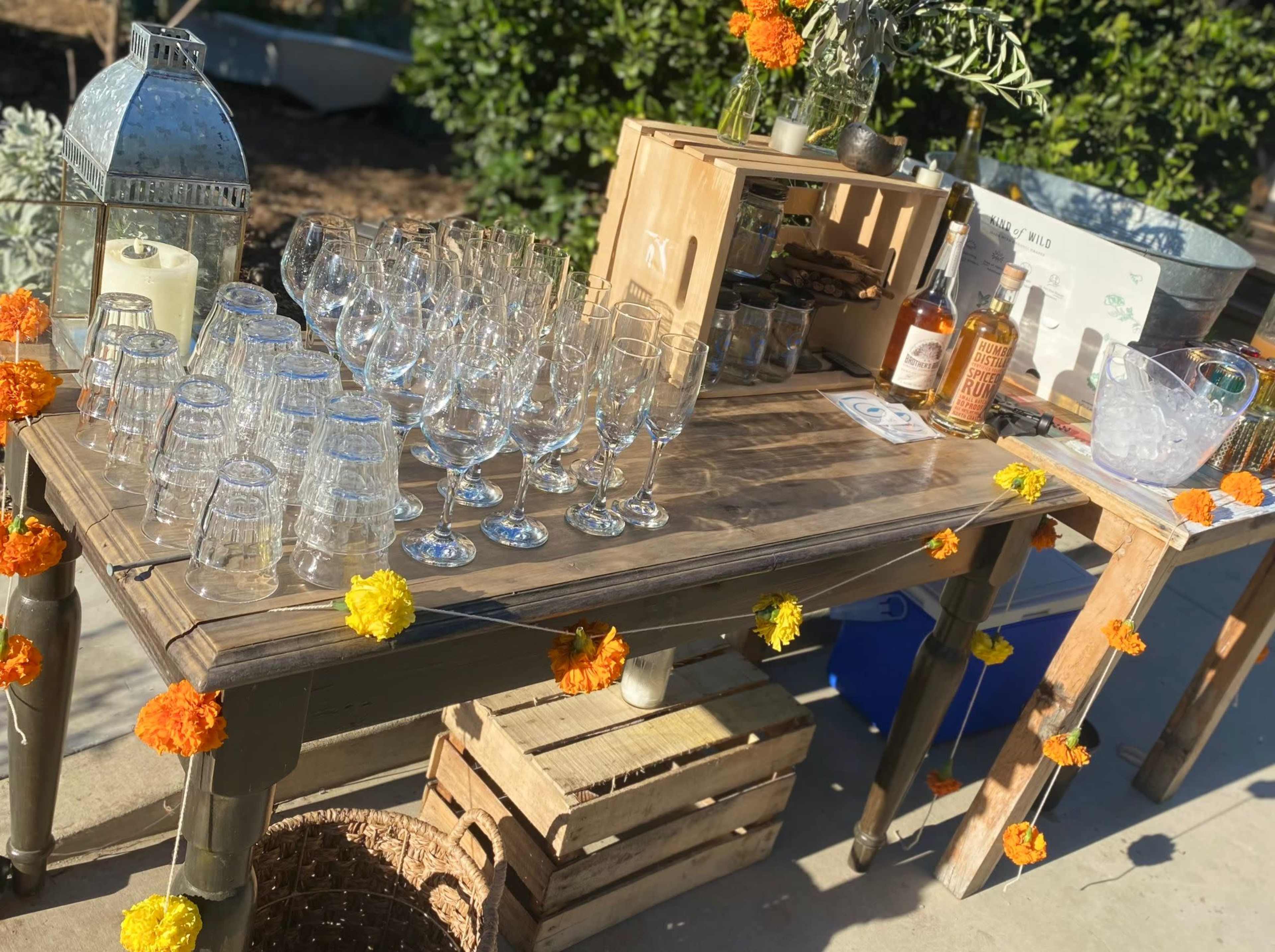 A wooden table is set up with an arrangement of clear glasses, bottles of whiskey, and decorative marigold flowers.