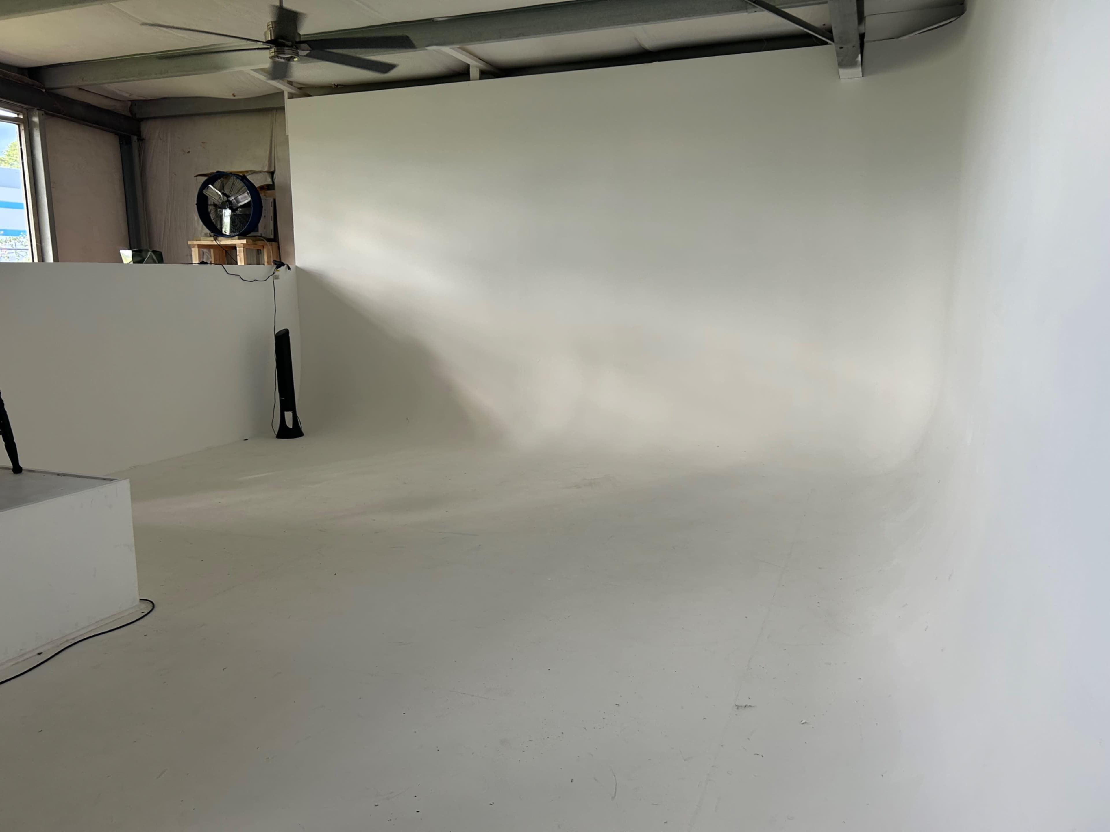 The image shows a spacious, empty studio with a smooth white backdrop and a fan in the corner.