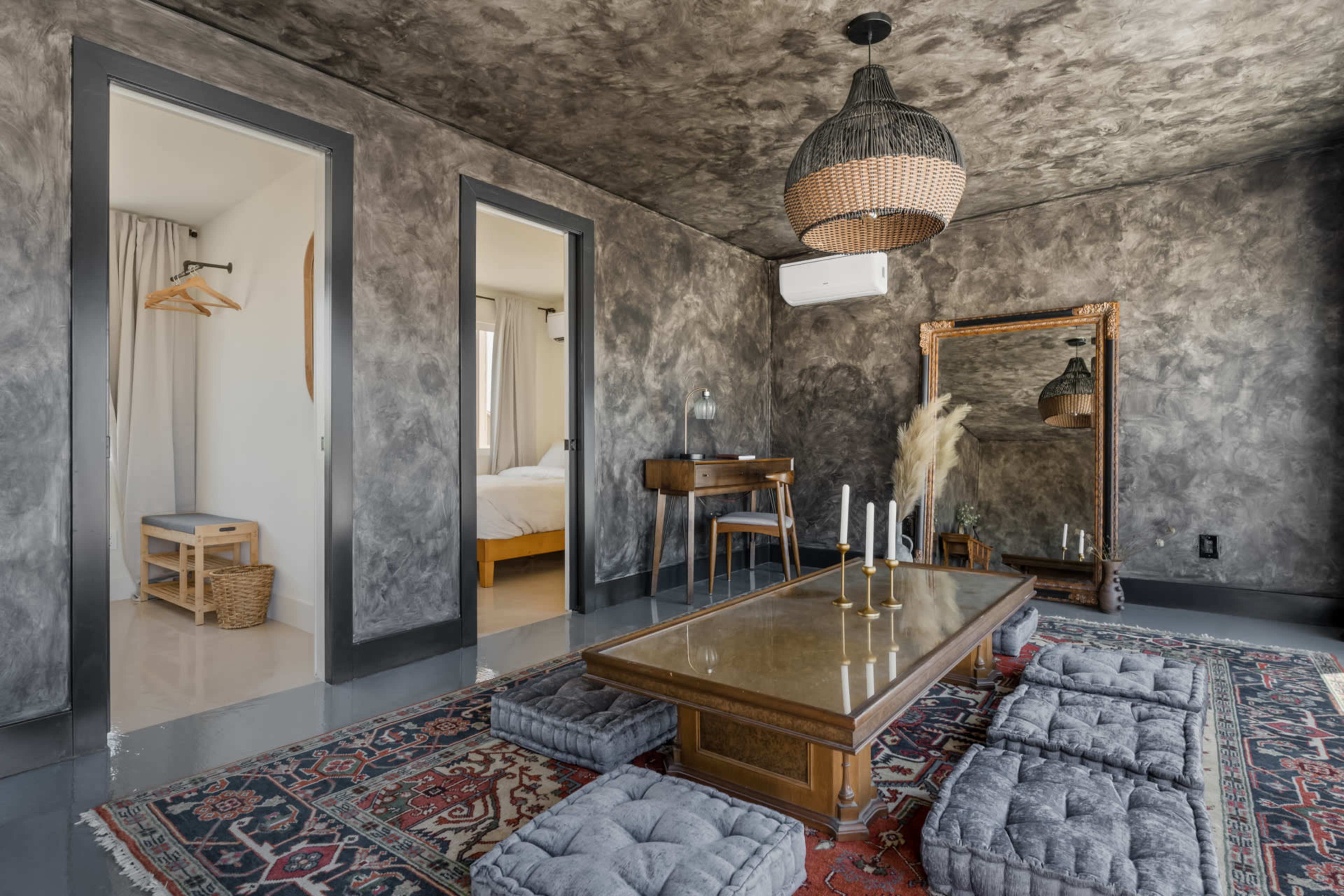The image shows a modern room with textured gray walls, a low wooden table surrounded by floor cushions, and a large mirror reflecting the space.