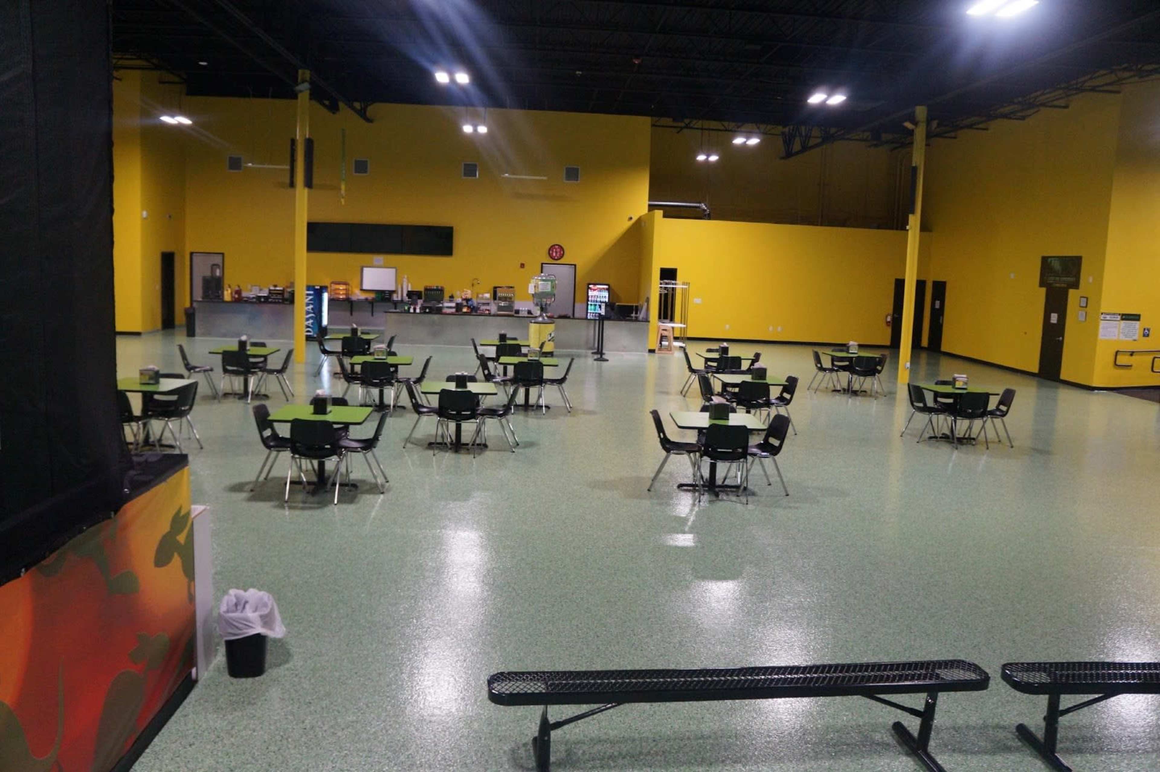 A large indoor space with multiple empty tables and chairs set up, surrounded by yellow walls and green flooring.
