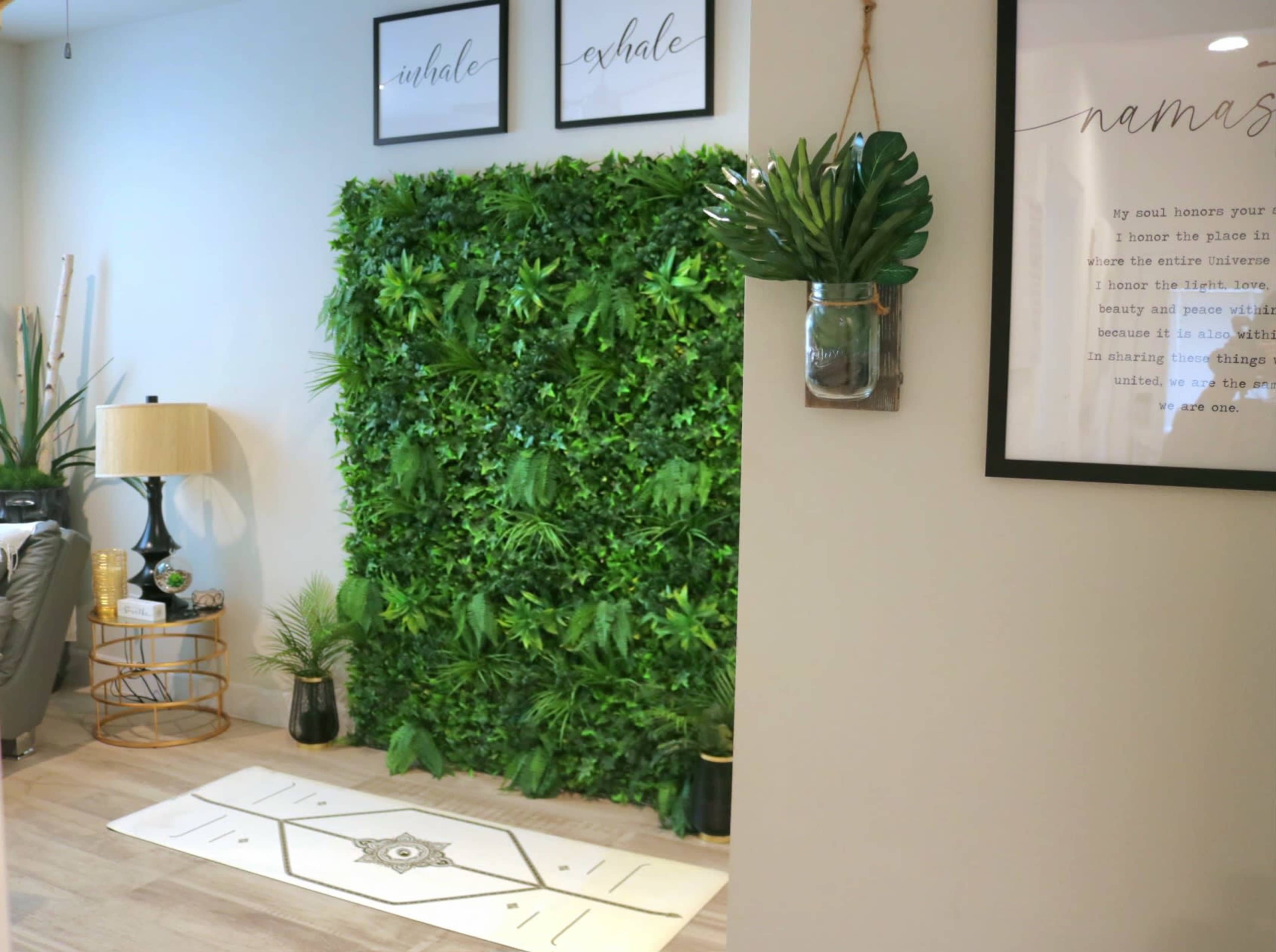 The image shows a modern interior space featuring a green plant wall, a lamp, decorative plants, and framed wall art that includes the words "inhale" and "exhale."