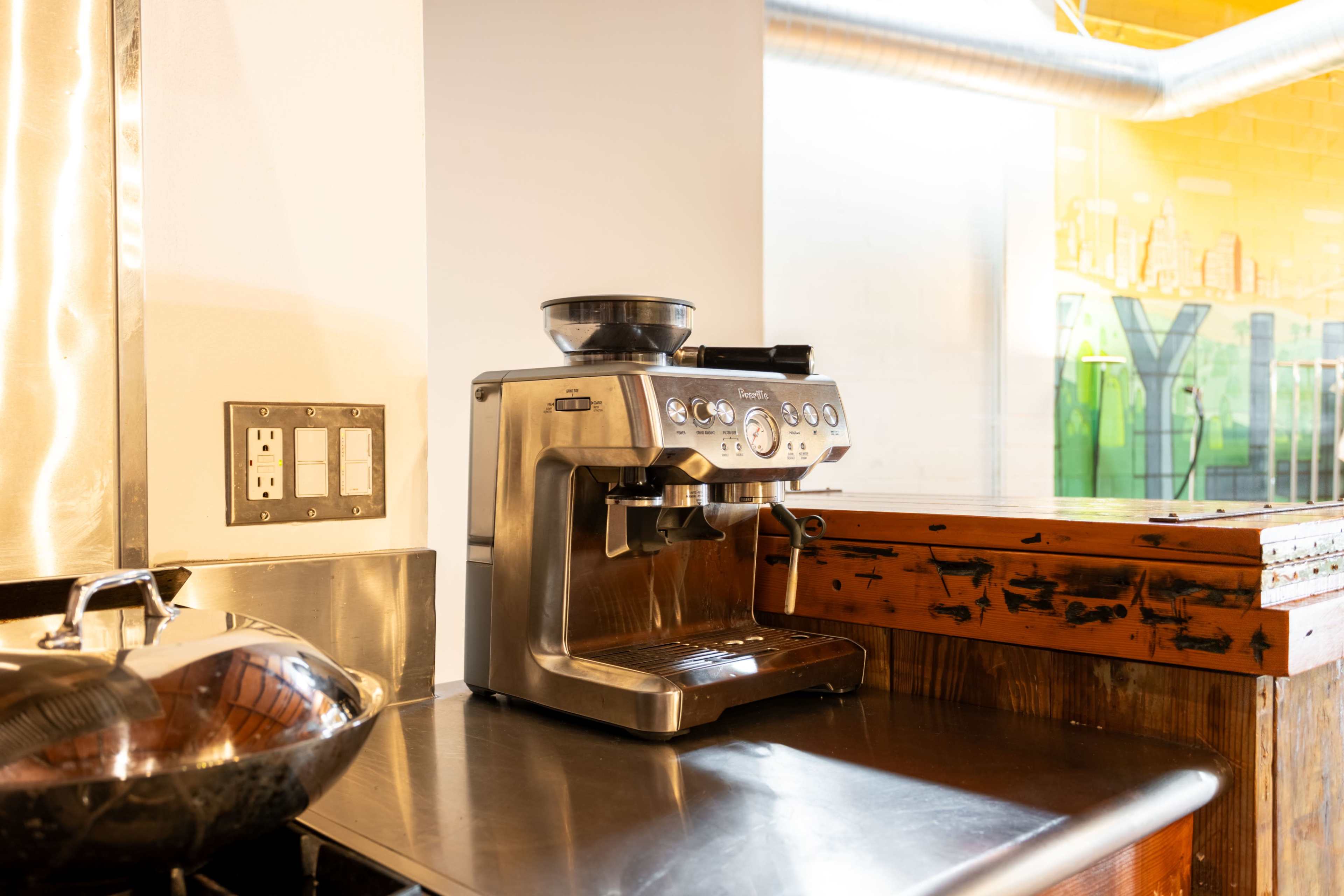 A stainless steel espresso machine is positioned on a wooden countertop next to a sink in a brightly lit space.