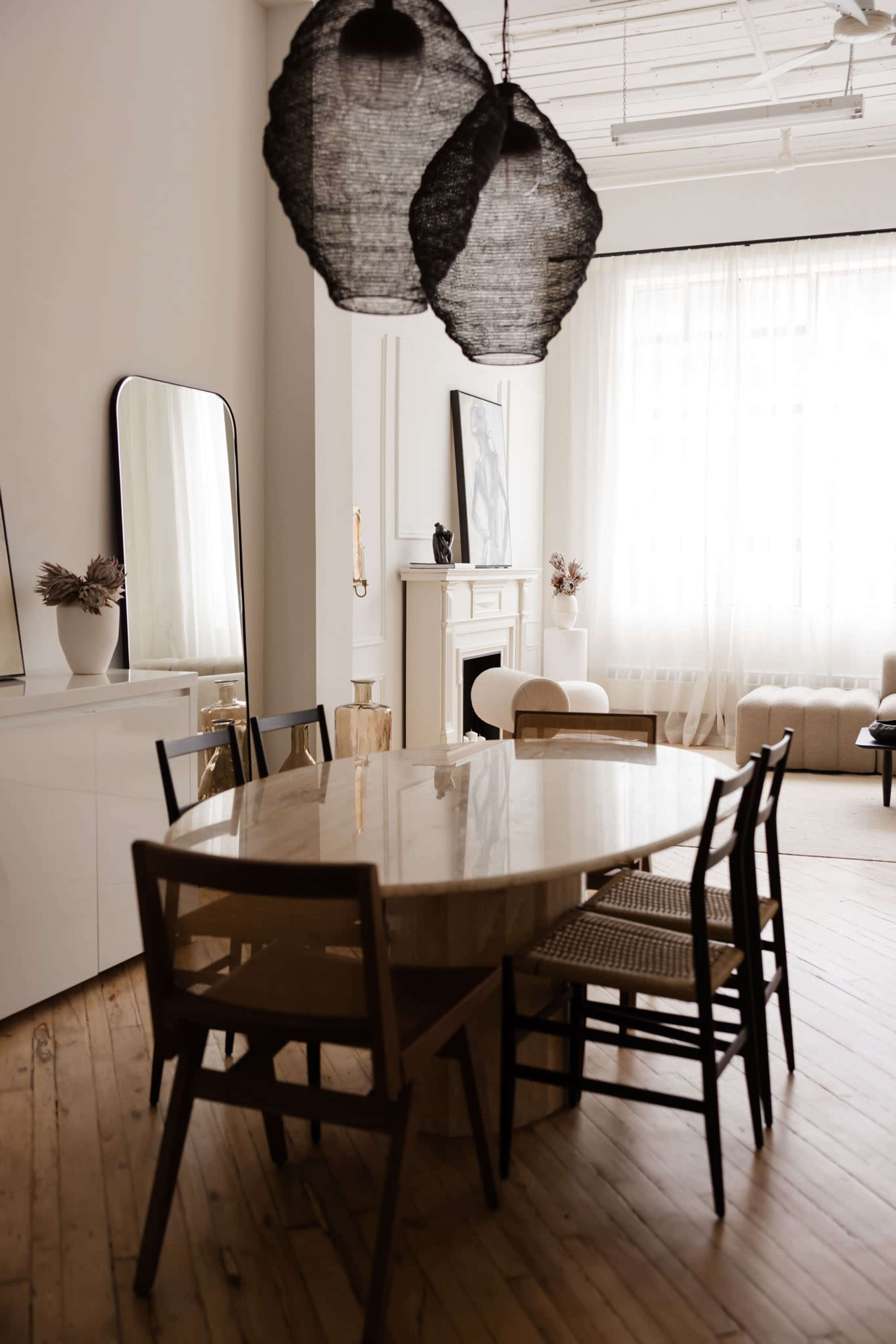 A modern dining area features a round marble table surrounded by wooden chairs, with prominent pendant lights overhead and large windows allowing natural light to fill the space.