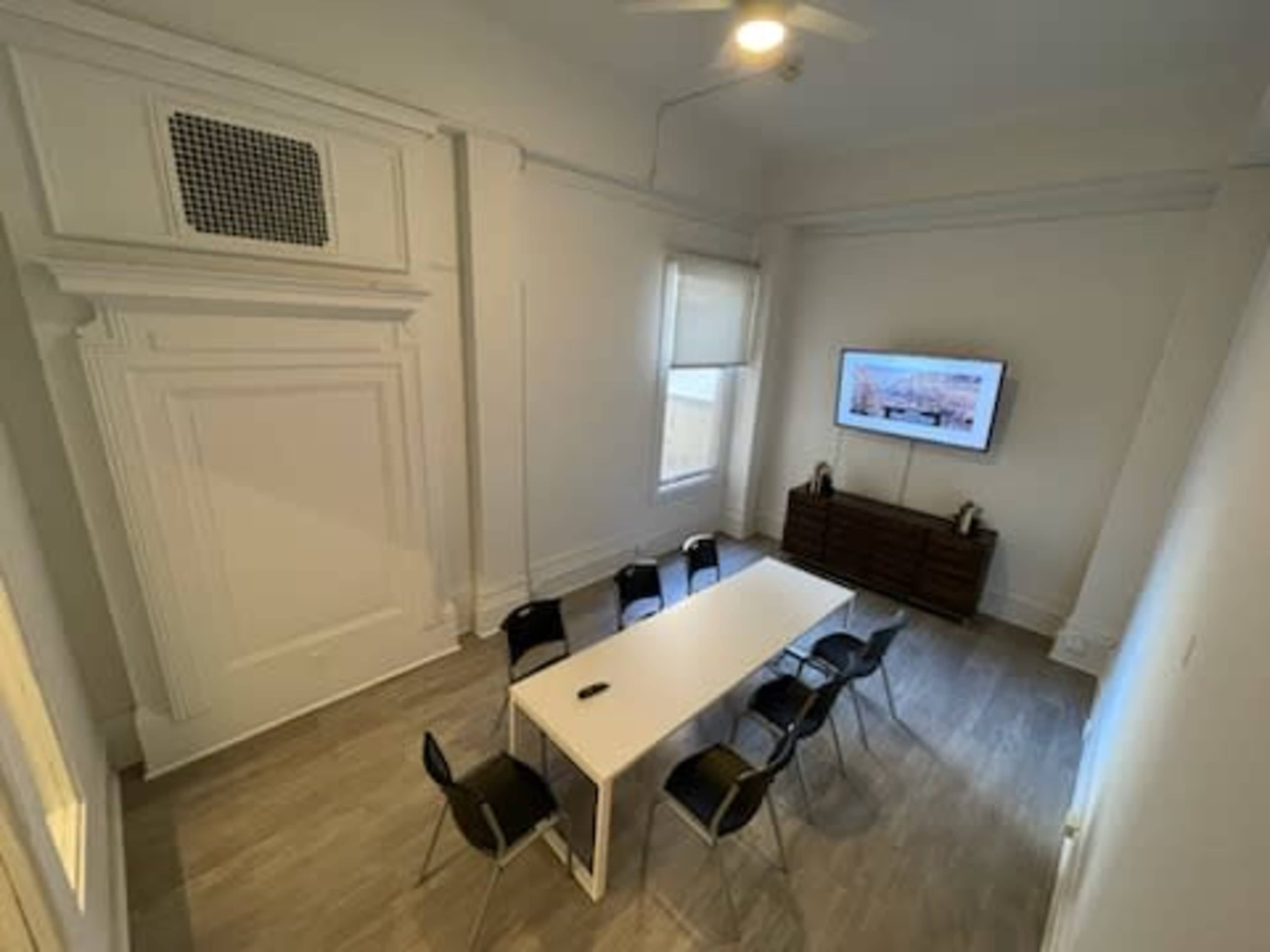 The image shows a conference room with a long white table surrounded by six black chairs, a wall-mounted television, and large windows allowing natural light.