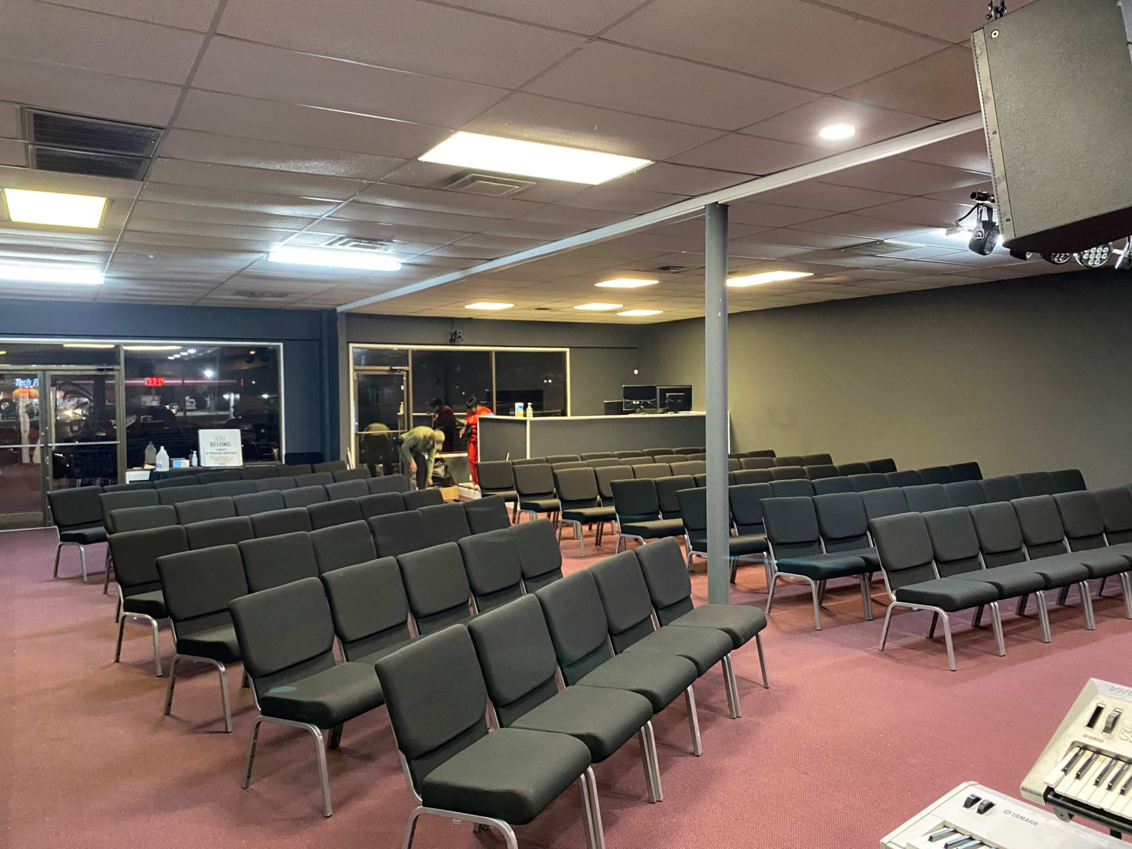 A spacious room with rows of black chairs set up for seating, featuring a stage area and sound equipment in the background.