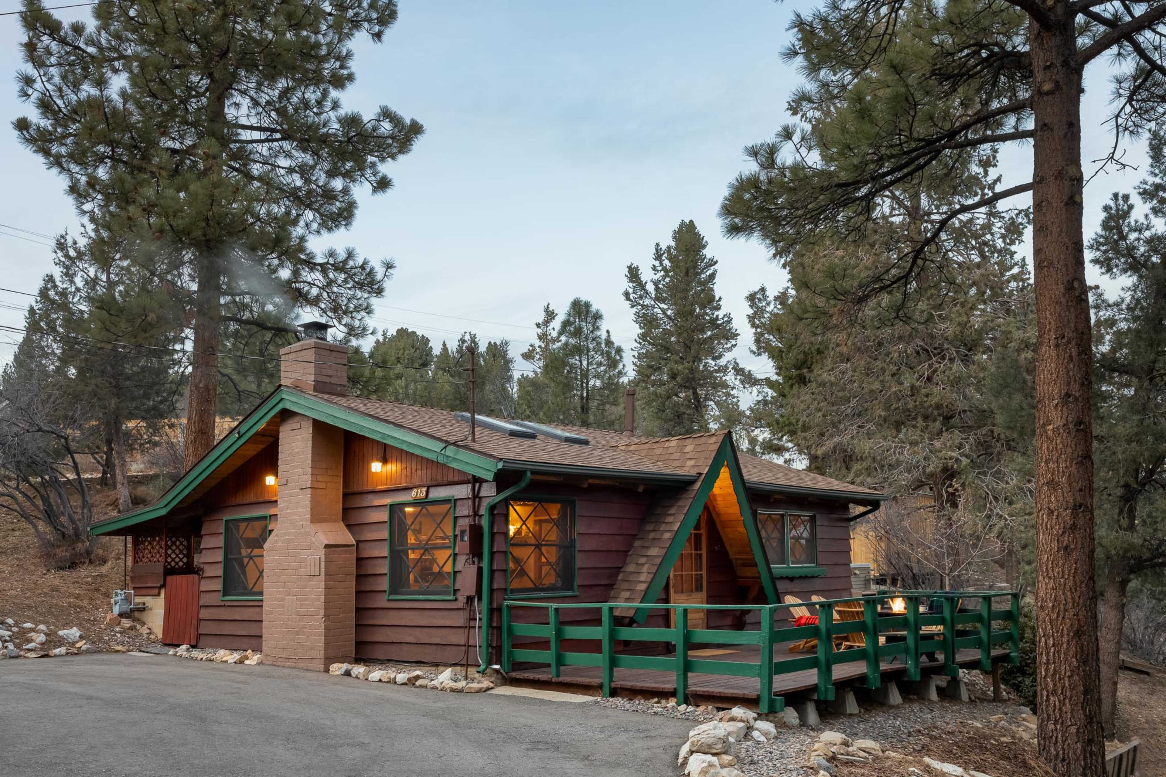 A brown cabin with green trim and a chimney sits among pine trees, along a gravel driveway.