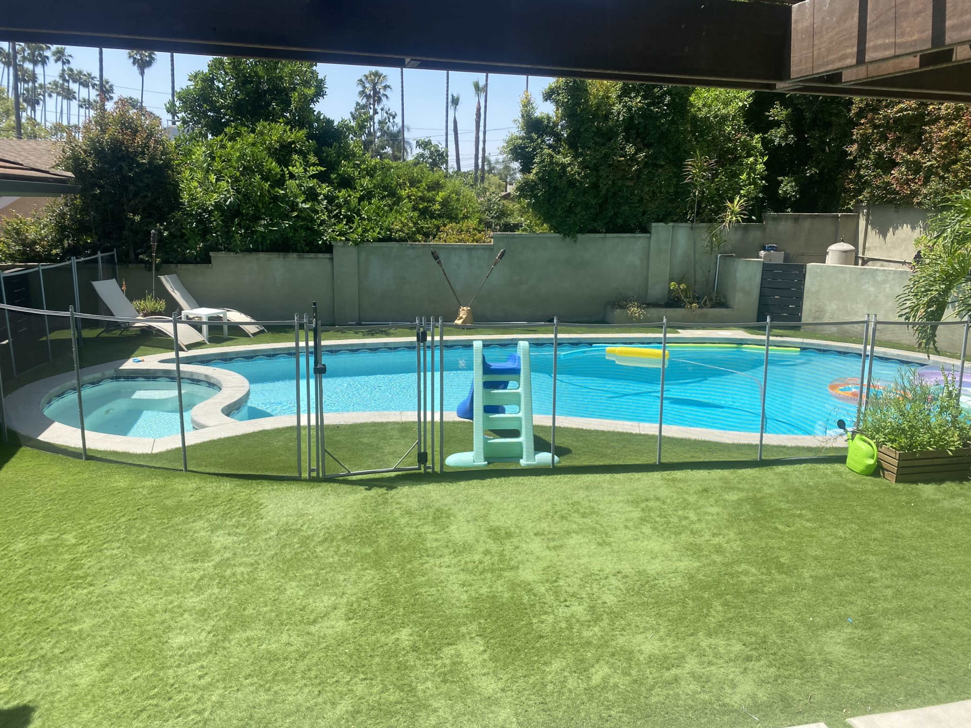 A rectangular swimming pool surrounded by a green lawn and enclosed by a safety fence, with lounge chairs and colorful pool floats nearby.