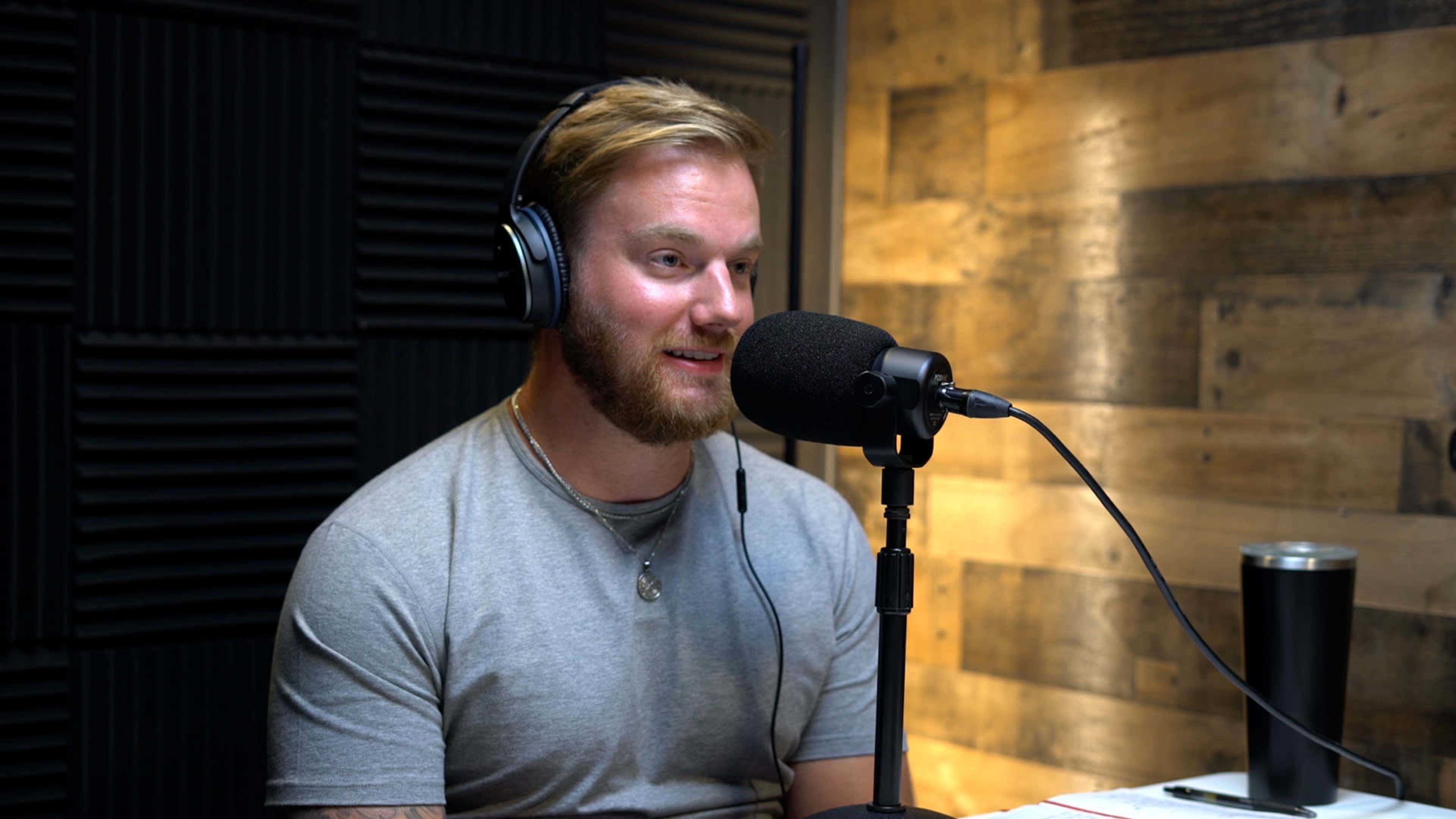 A man with a beard sits in a recording studio, speaking into a microphone while wearing headphones.