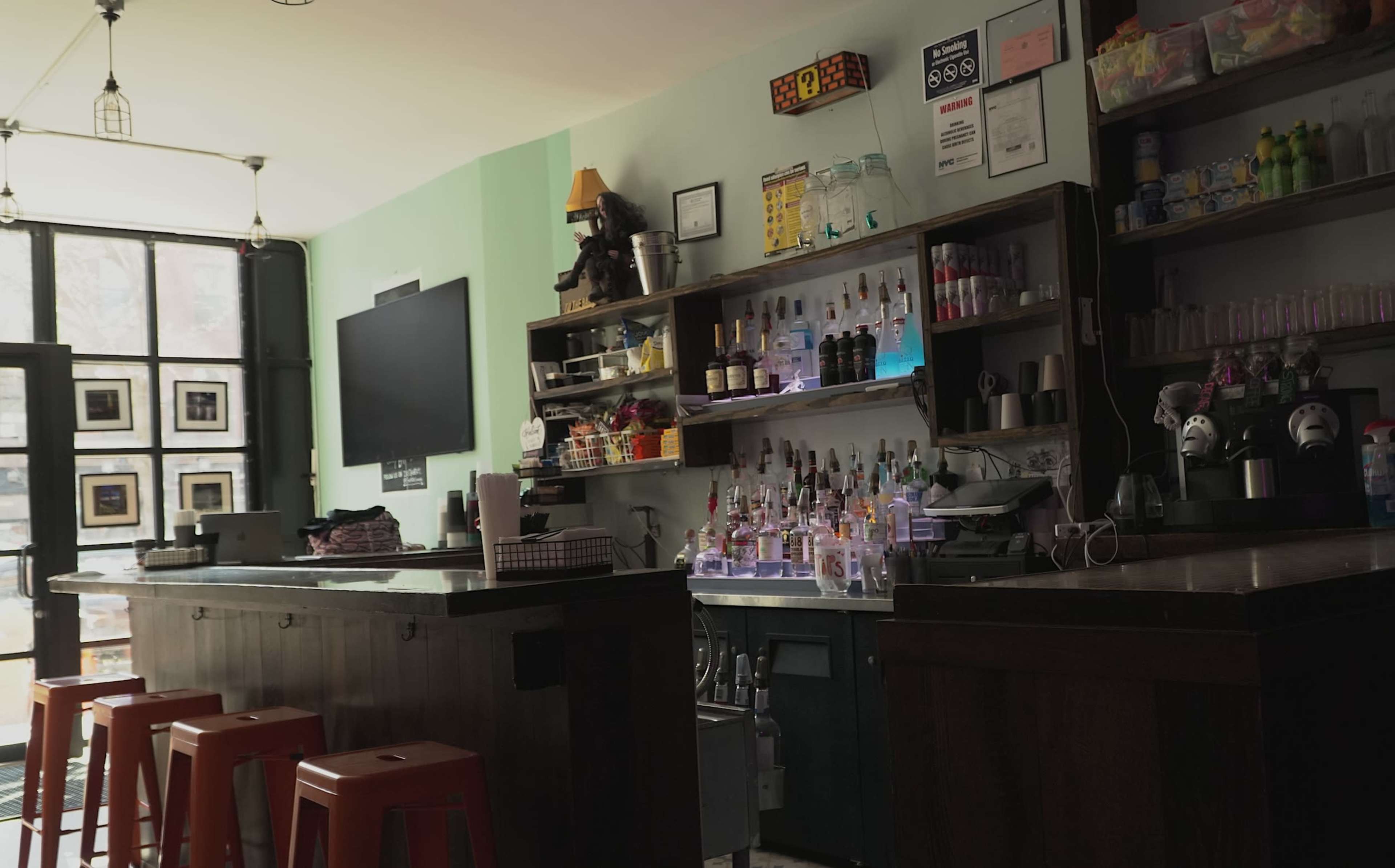 The image shows an empty bar area with a wooden counter, colorful stools, and shelves filled with various bottles and glassware.