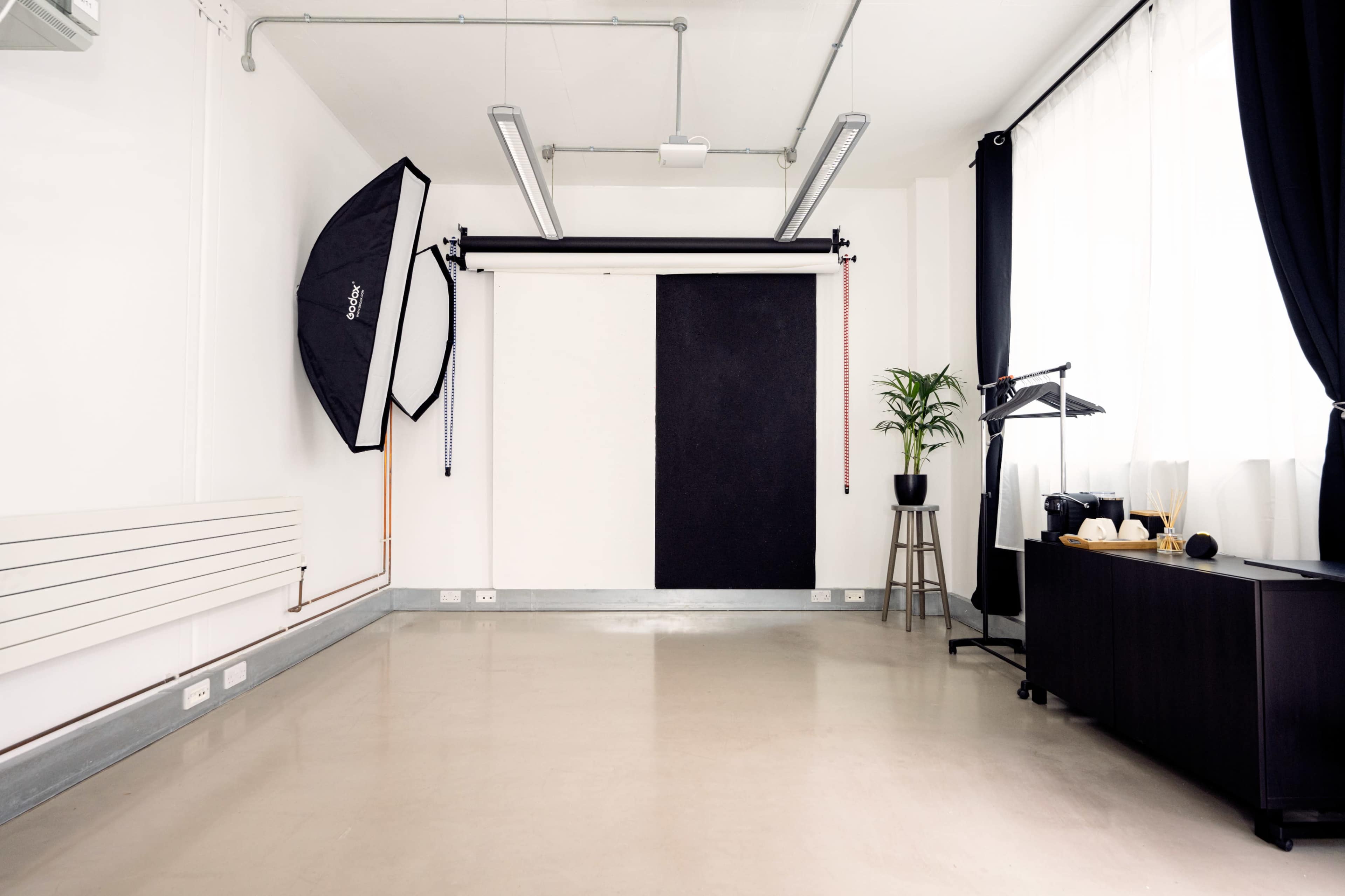 A photography studio features a backdrop, softbox light, a potted plant, and minimalist furniture against white walls.