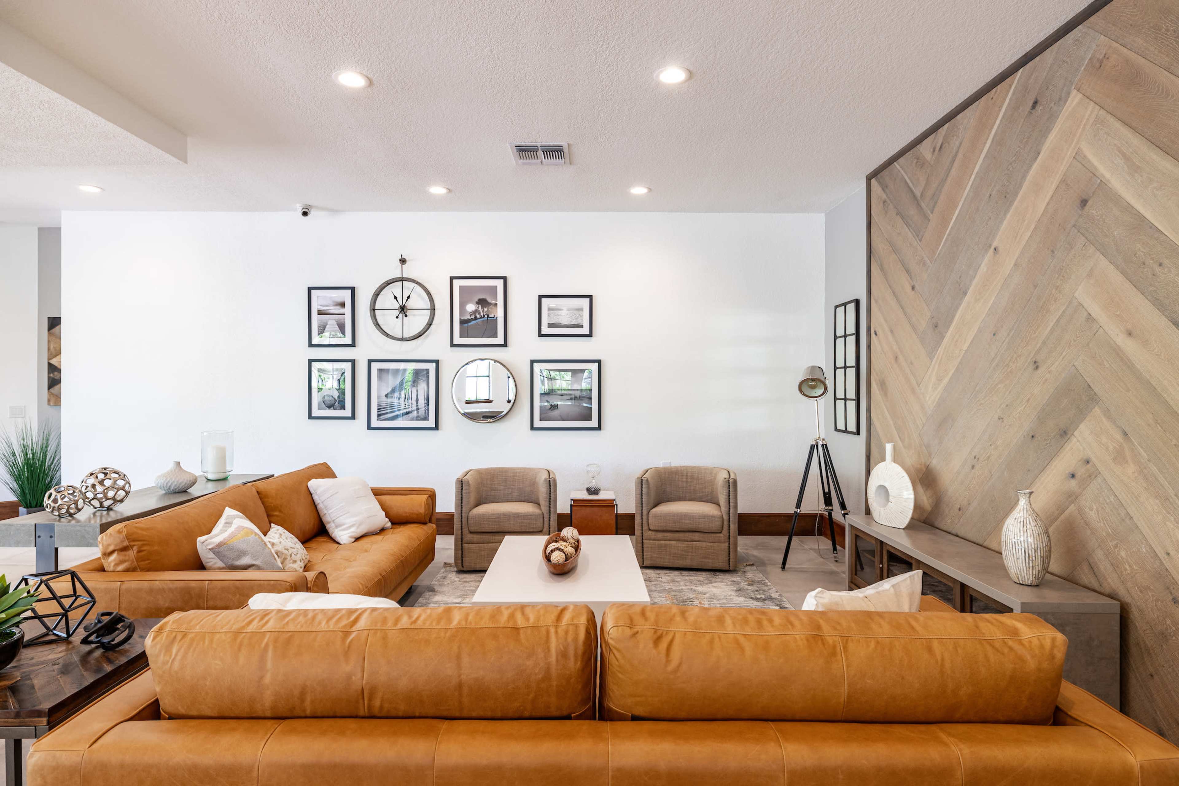 A living room features two brown leather sofas, three beige upholstered chairs, a wooden coffee table, and a gallery of framed photos on the white wall.
