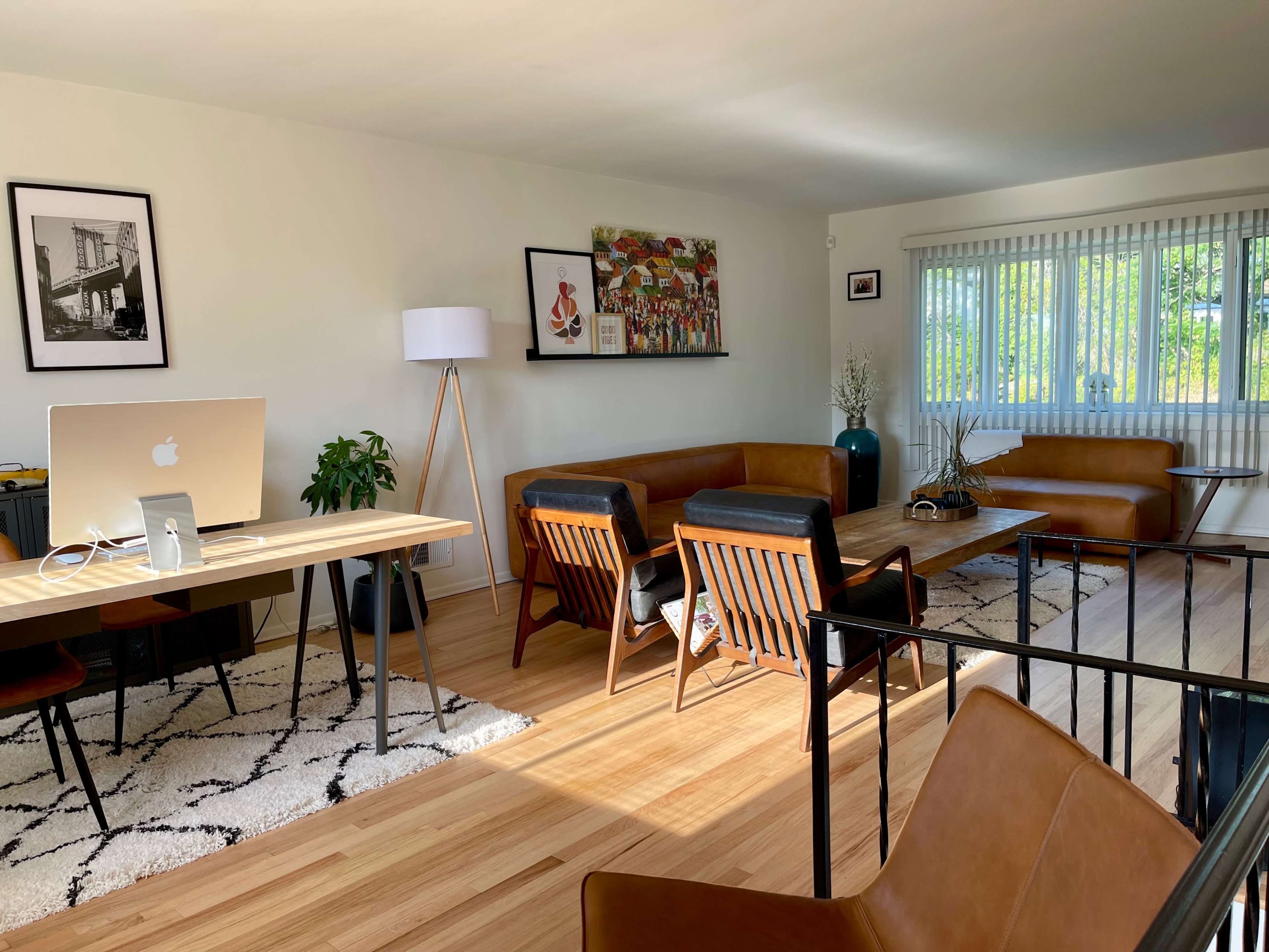 A modern living room features a desk with a computer, brown leather sofas, wooden chairs, and large windows allowing natural light to fill the space.