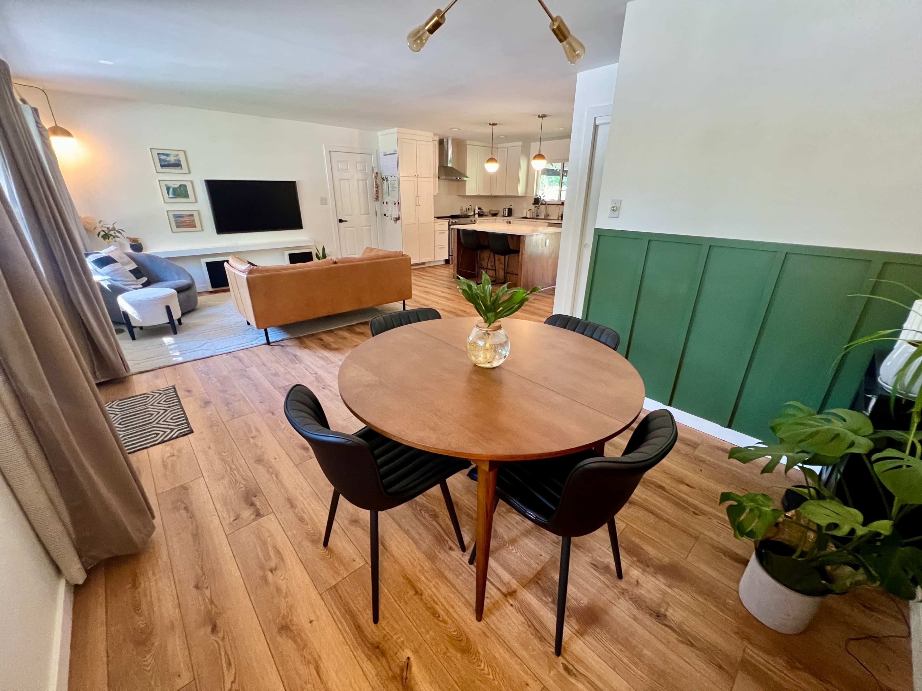 A dining area features a round wooden table surrounded by black chairs, with a living room visible in the background.