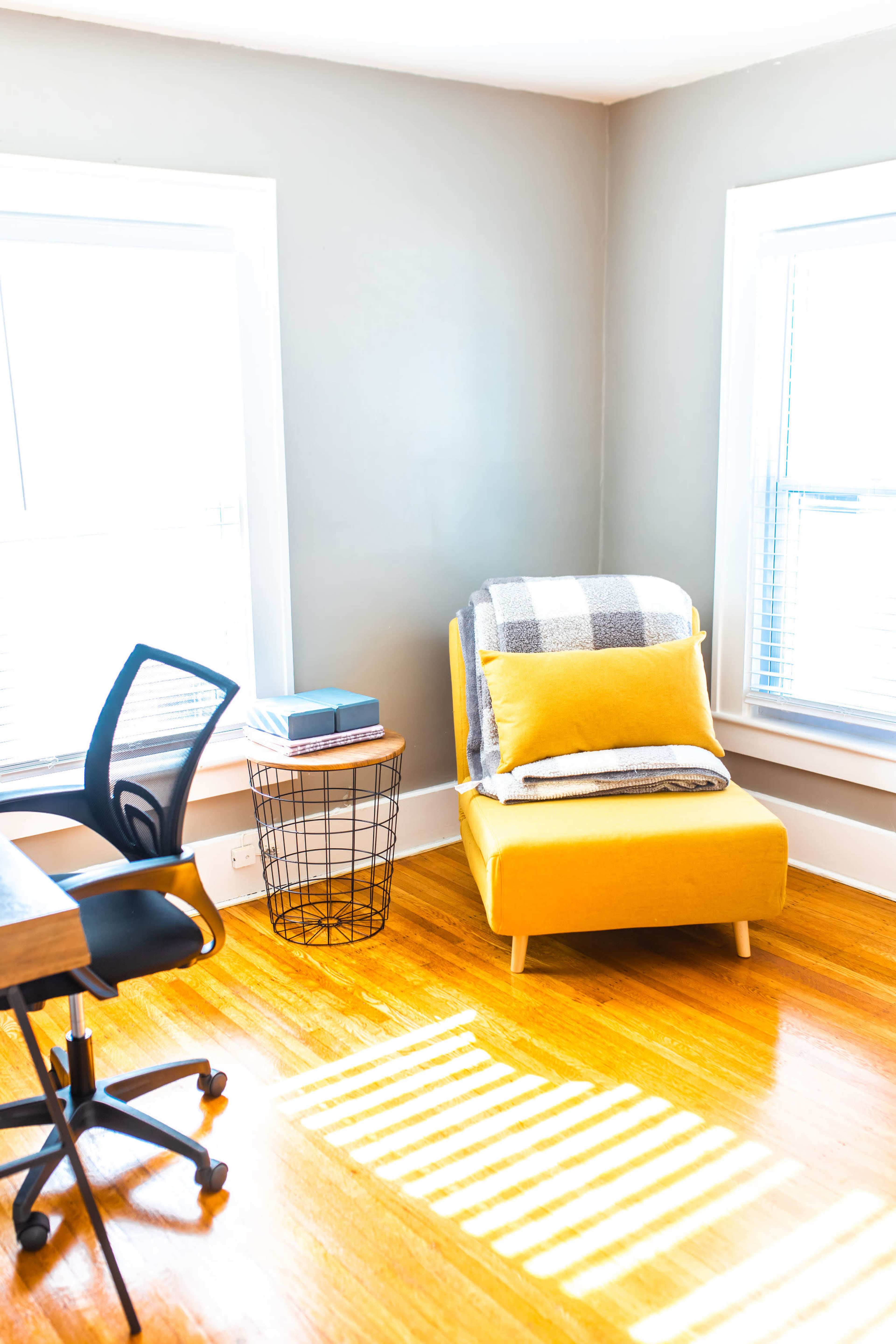 A bright corner of a room featuring a yellow chair with a blanket, a side table with a book, and a black office chair positioned near a wooden desk.