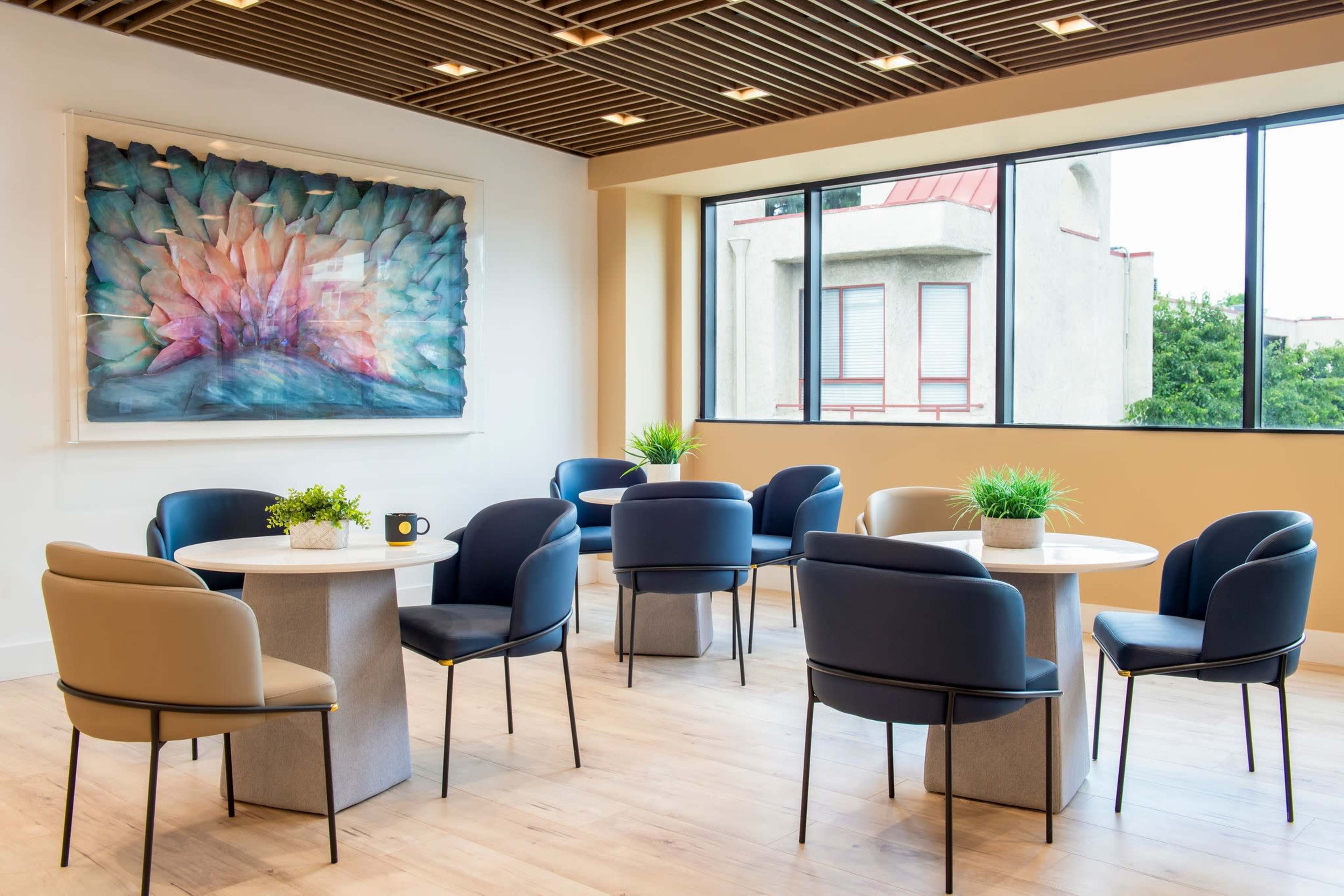 The image shows a modern conference area with round tables, chairs in various colors, and a large colorful artwork on the wall beside a window.