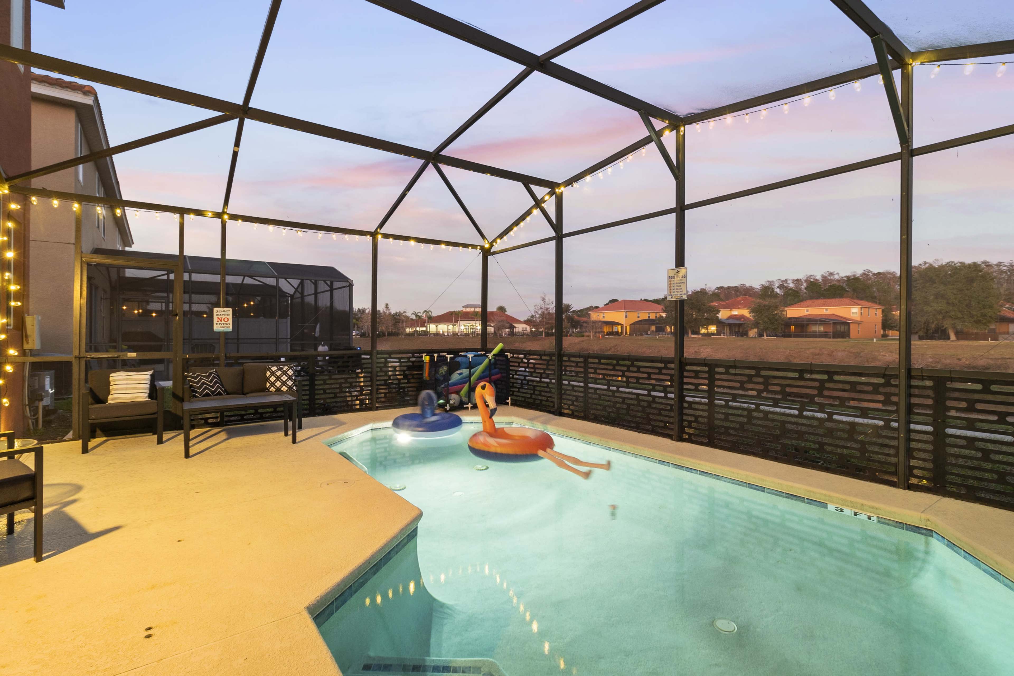 The image shows a screened-in pool area with colorful floaties, outdoor seating, and residential buildings in the background.