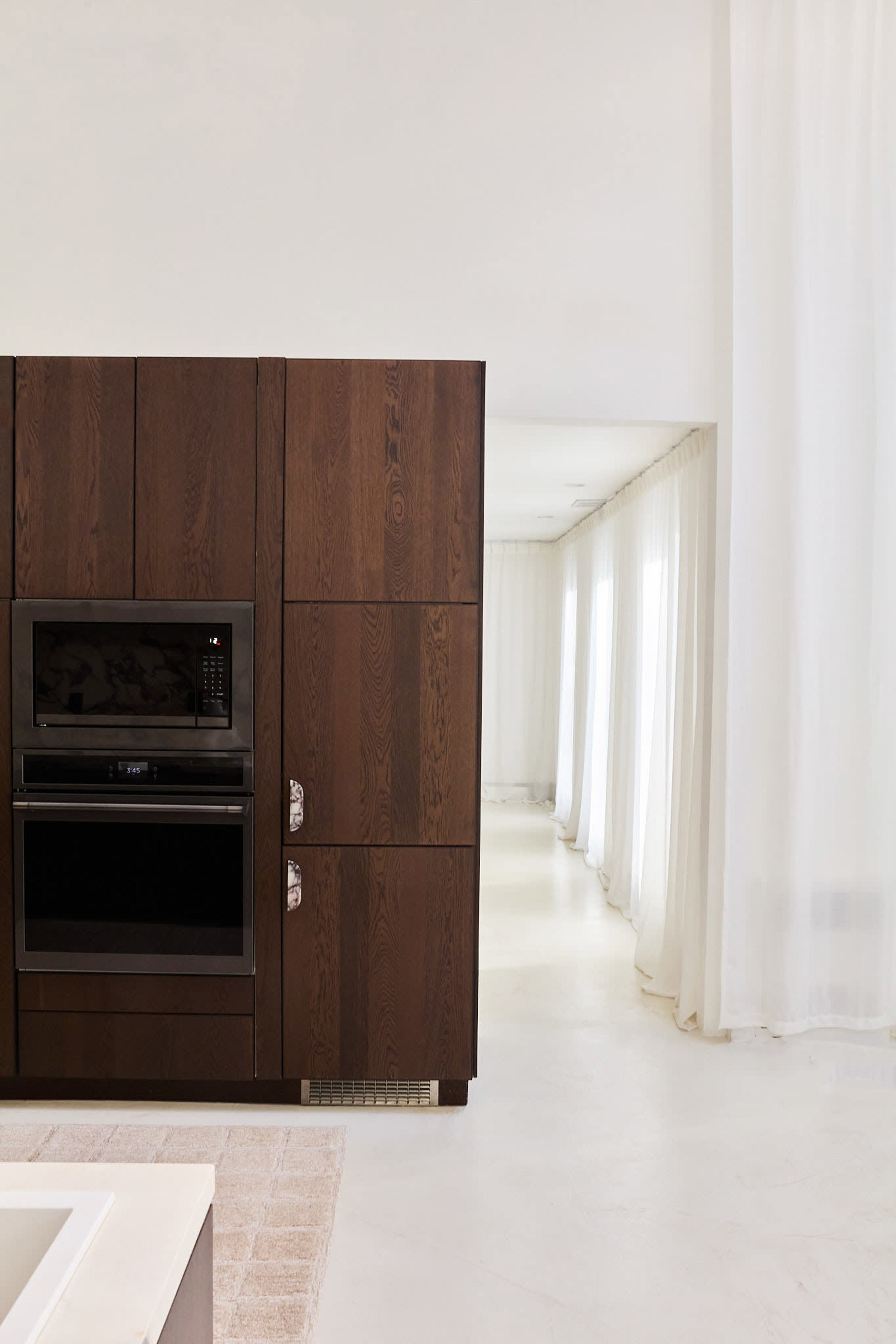 The image shows a modern kitchen with a dark wooden cabinet containing an oven and a long corridor lined with sheer white curtains.