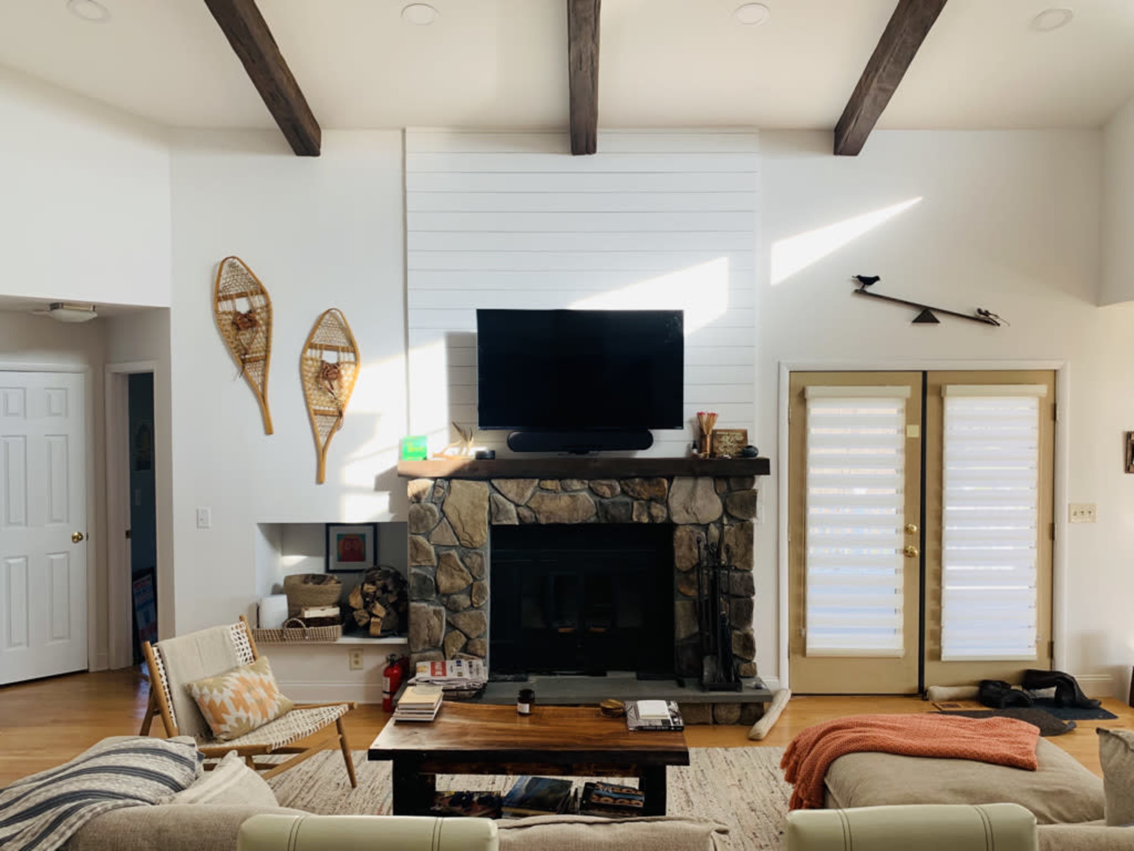 A cozy living room with a stone fireplace, a mounted television above it, and large glass doors leading outside, complemented by wooden beams on the ceiling.