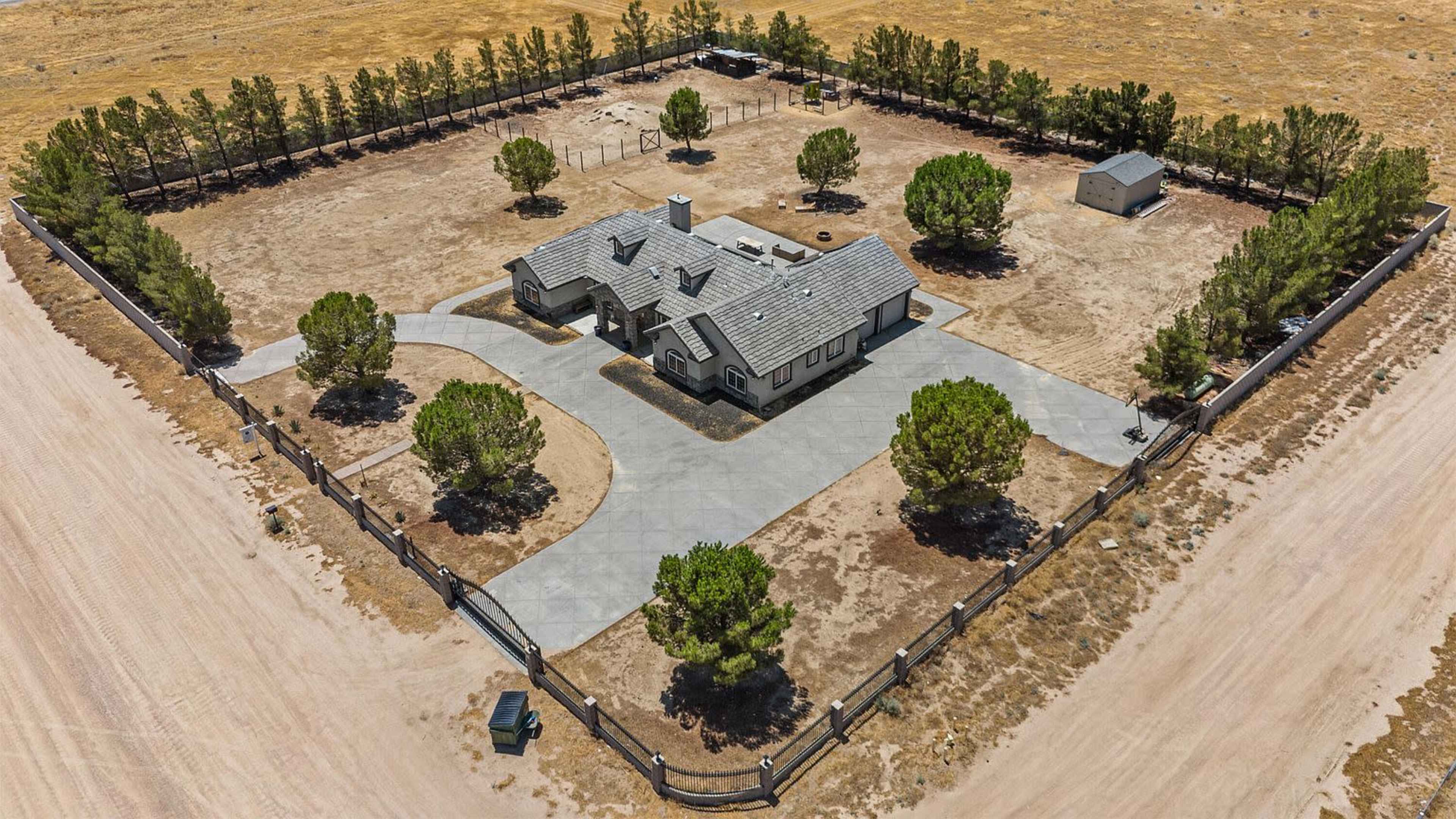 The image depicts a spacious single-story house surrounded by a gravel driveway and a fenced yard with several trees and a small outbuilding.