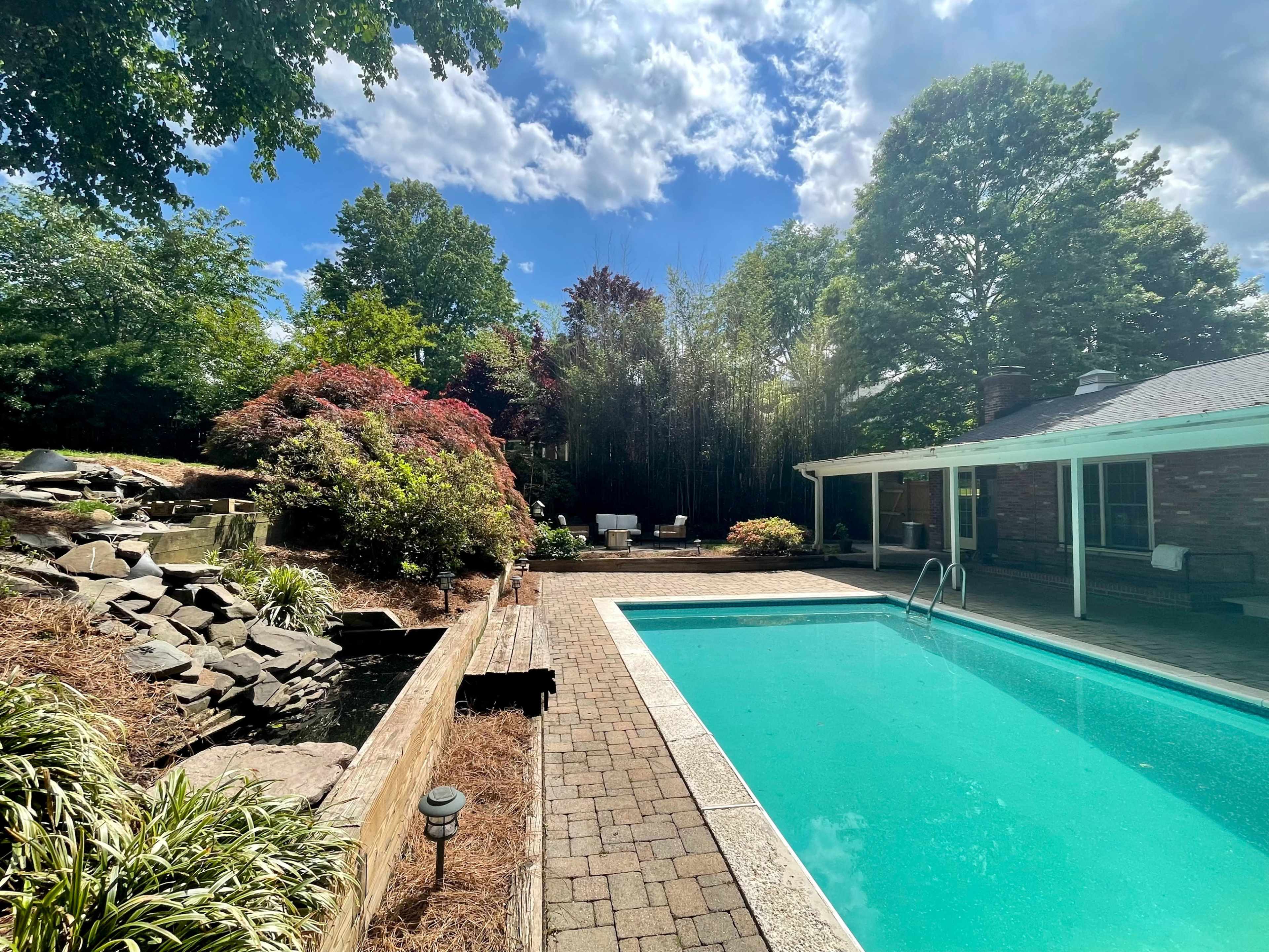 A clear swimming pool surrounded by a stone patio, garden beds, and a small waterfall features in a residential backyard.