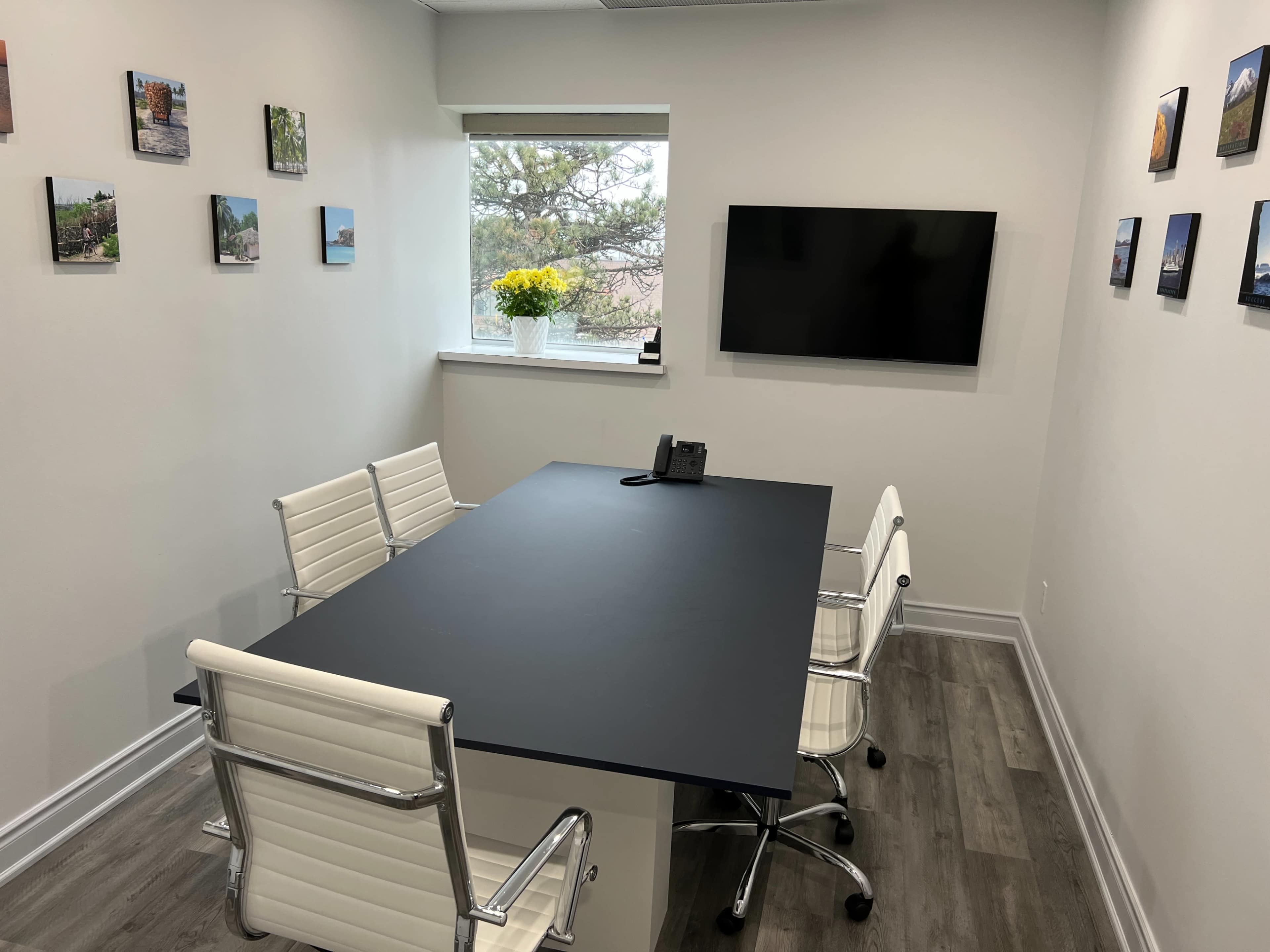 A conference room features a black table, four white chairs, a phone, and a television mounted on the wall, with framed photos arranged around the room and a window with flowers.