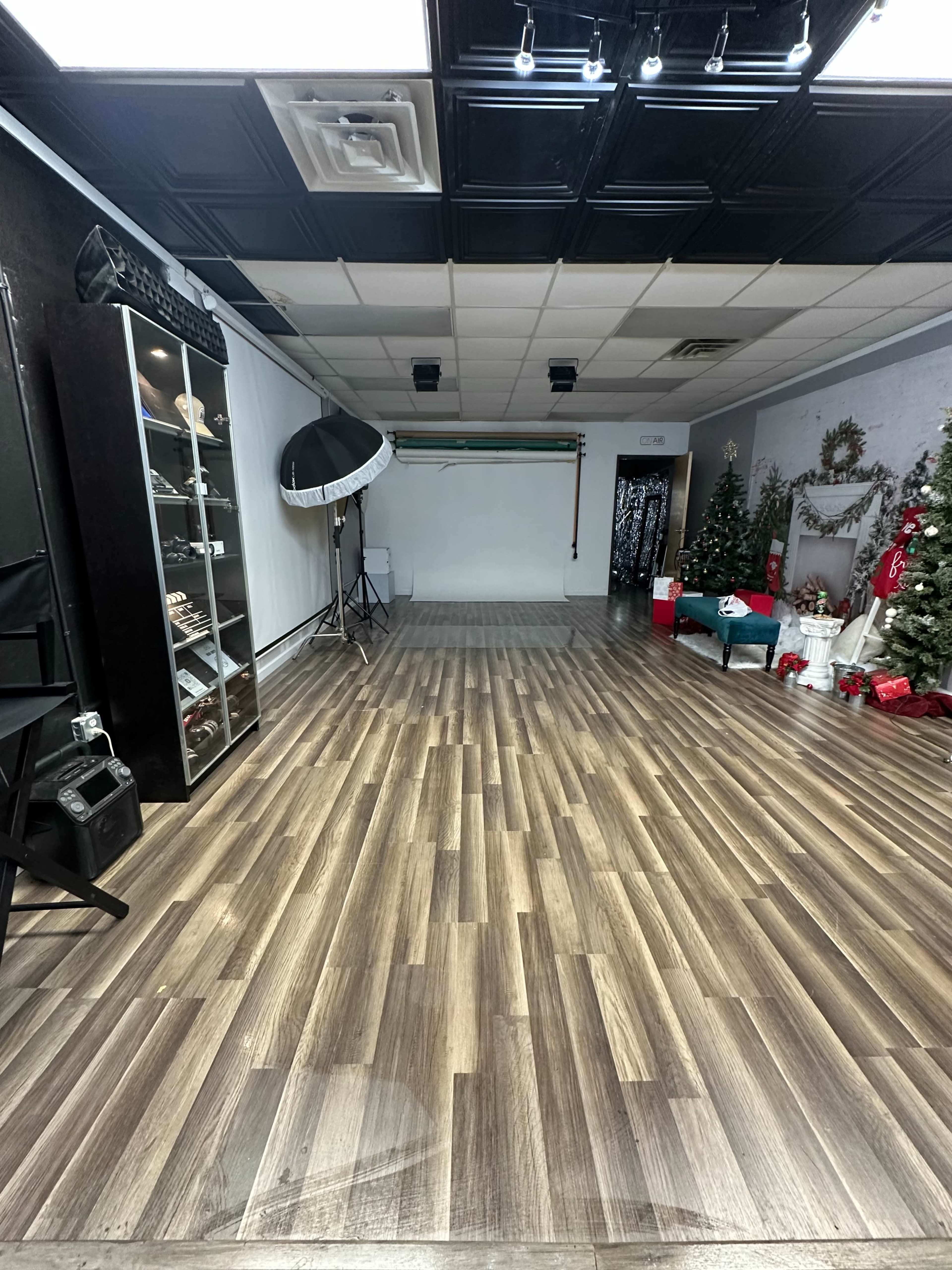 The image shows a photography studio with wooden flooring, a backdrop stand, a large light source umbrella, and decorated areas featuring Christmas trees and holiday décor along the walls.