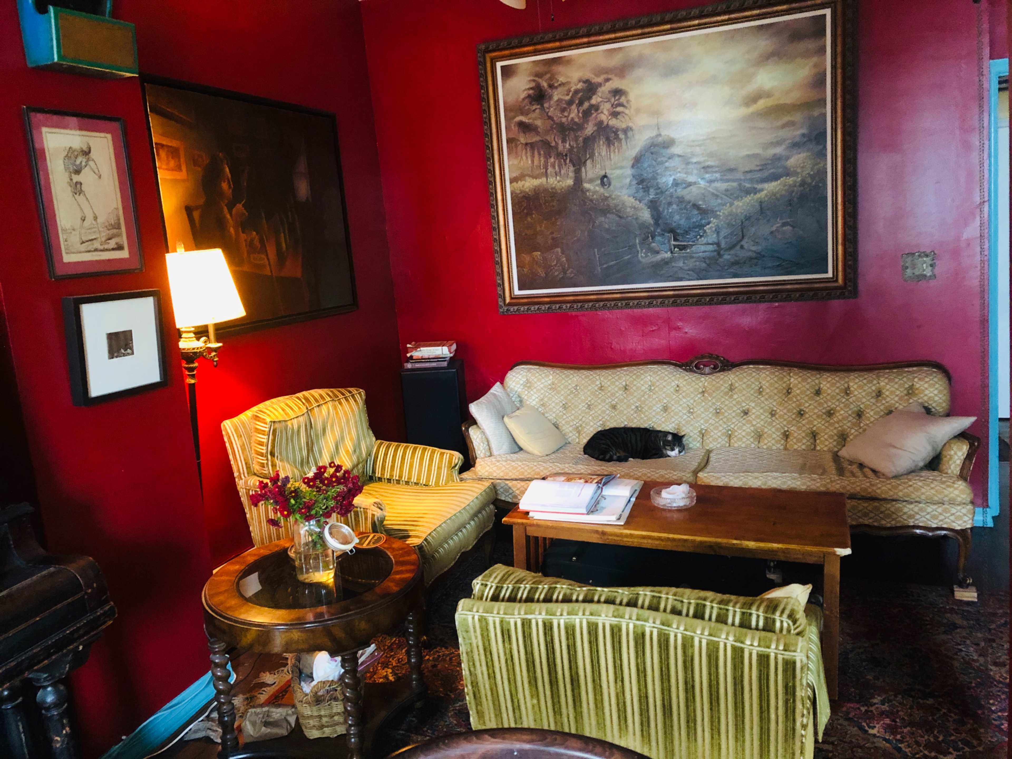The scene features a cozy living room with a red-painted wall, a vintage sofa and armchairs, a coffee table adorned with flowers and books, and a lamp providing warm light.