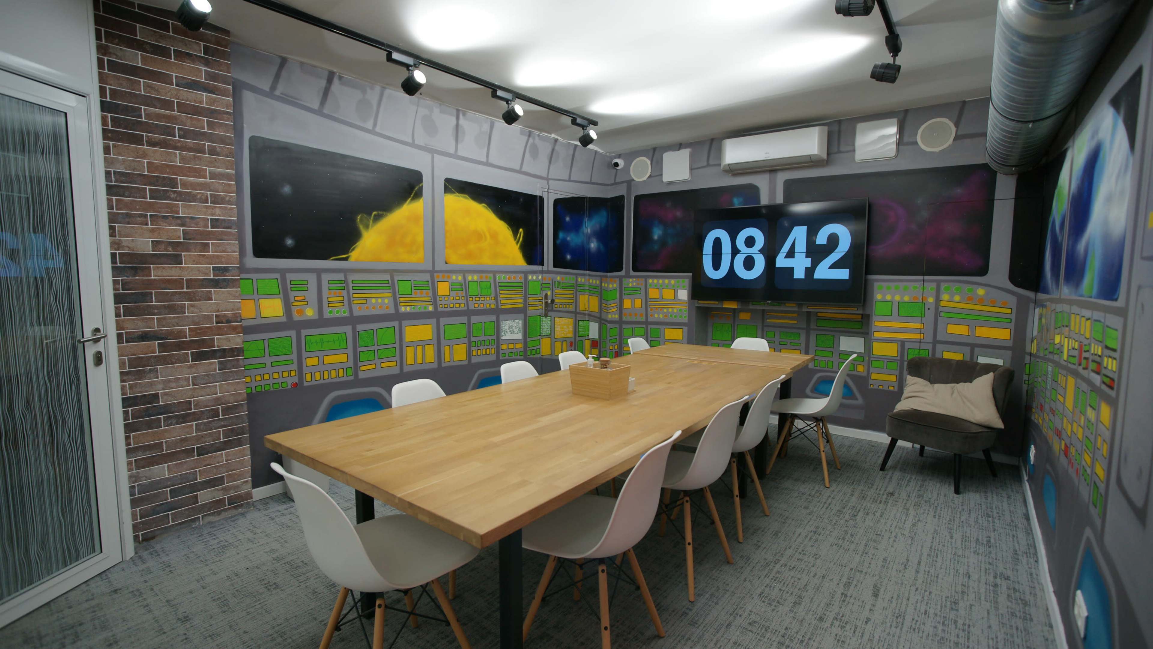 A modern meeting room features a large wooden table surrounded by white chairs, with vibrant wall murals depicting a control center and a digital clock displaying the time.
