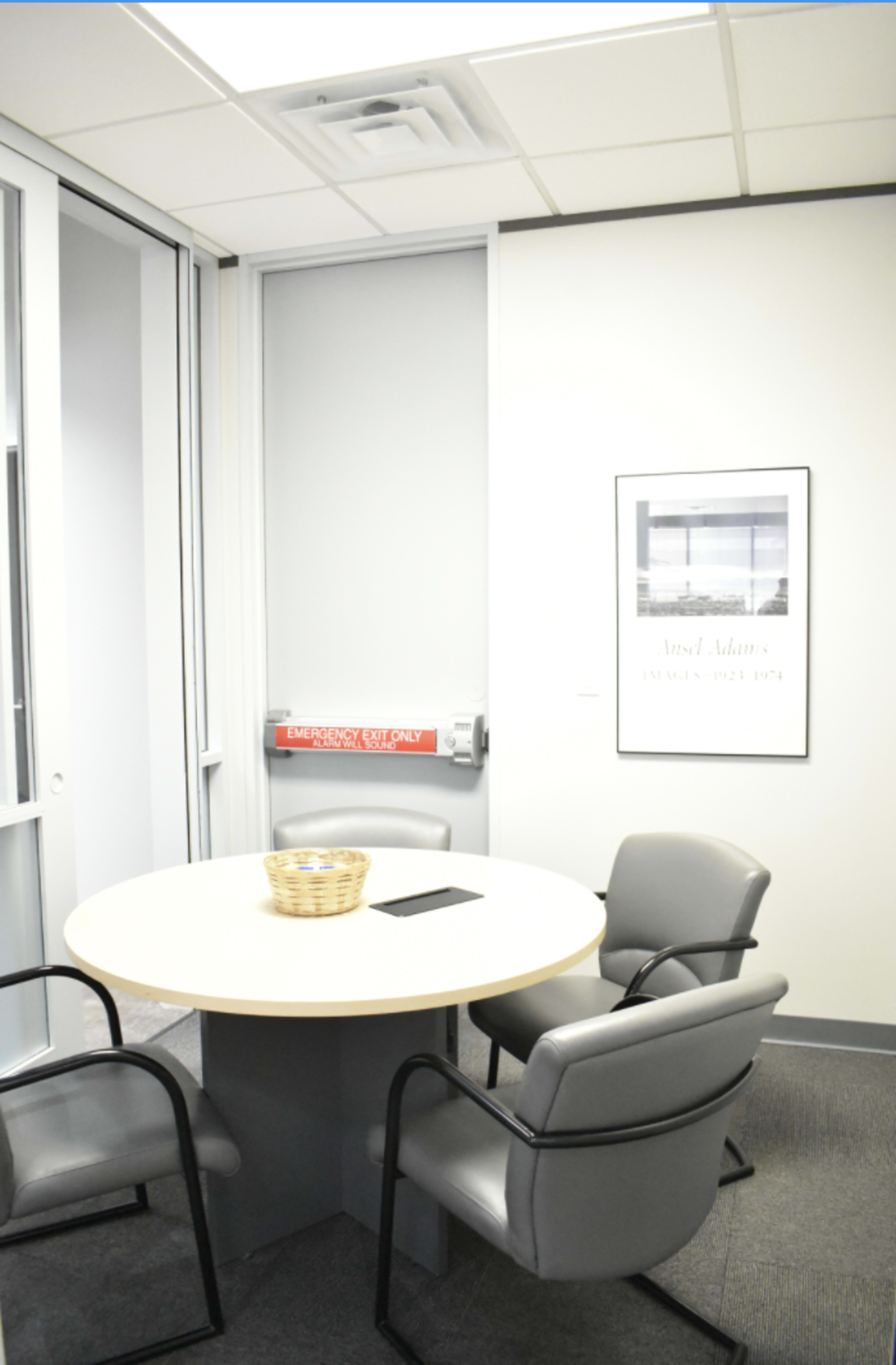 A small conference room features a round table surrounded by four chairs, with a fire extinguisher mounted on the wall and a framed poster nearby.