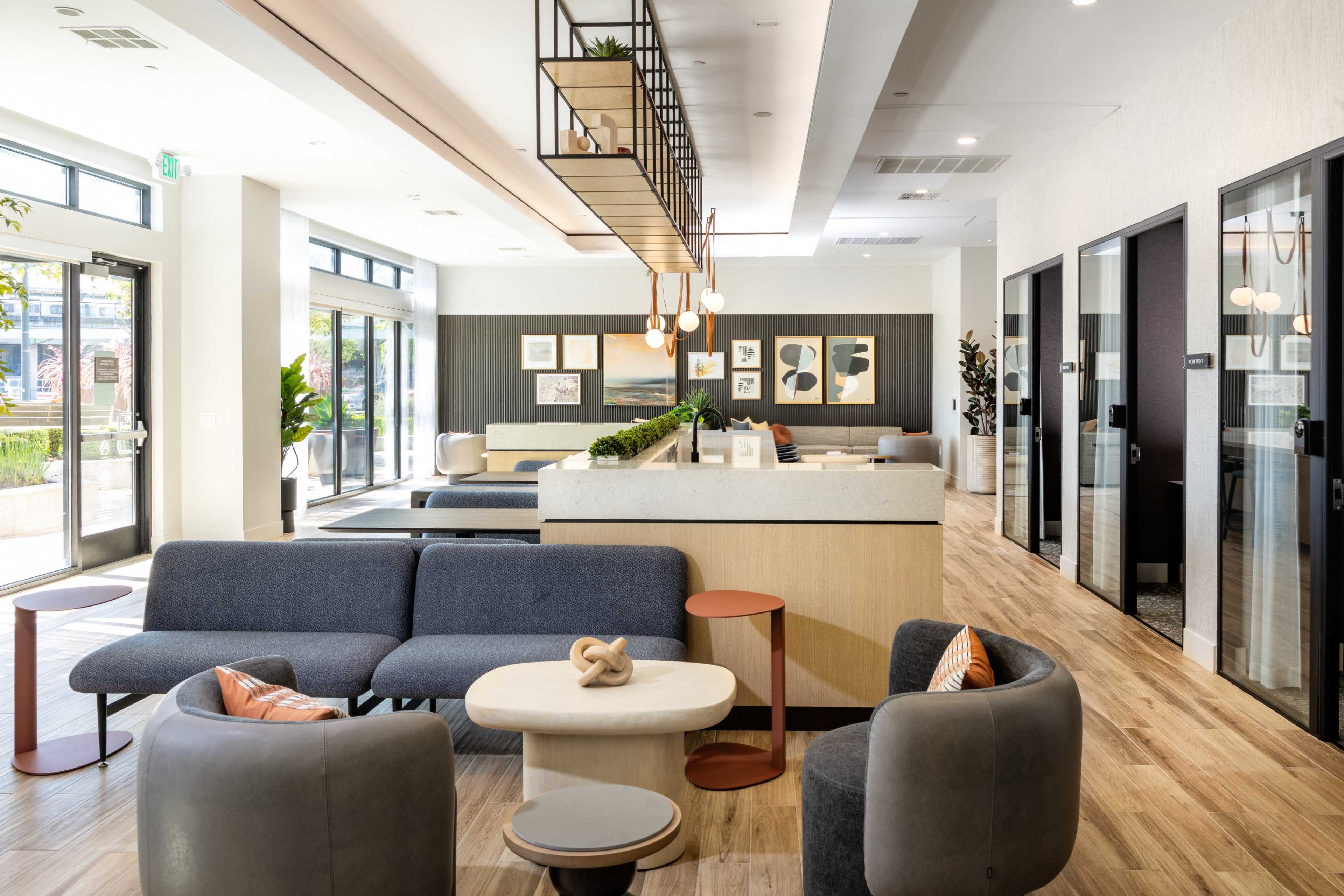 The image shows a modern lobby area featuring gray sofas, a circular coffee table, and a reception desk with decorative plants and artwork on the walls.