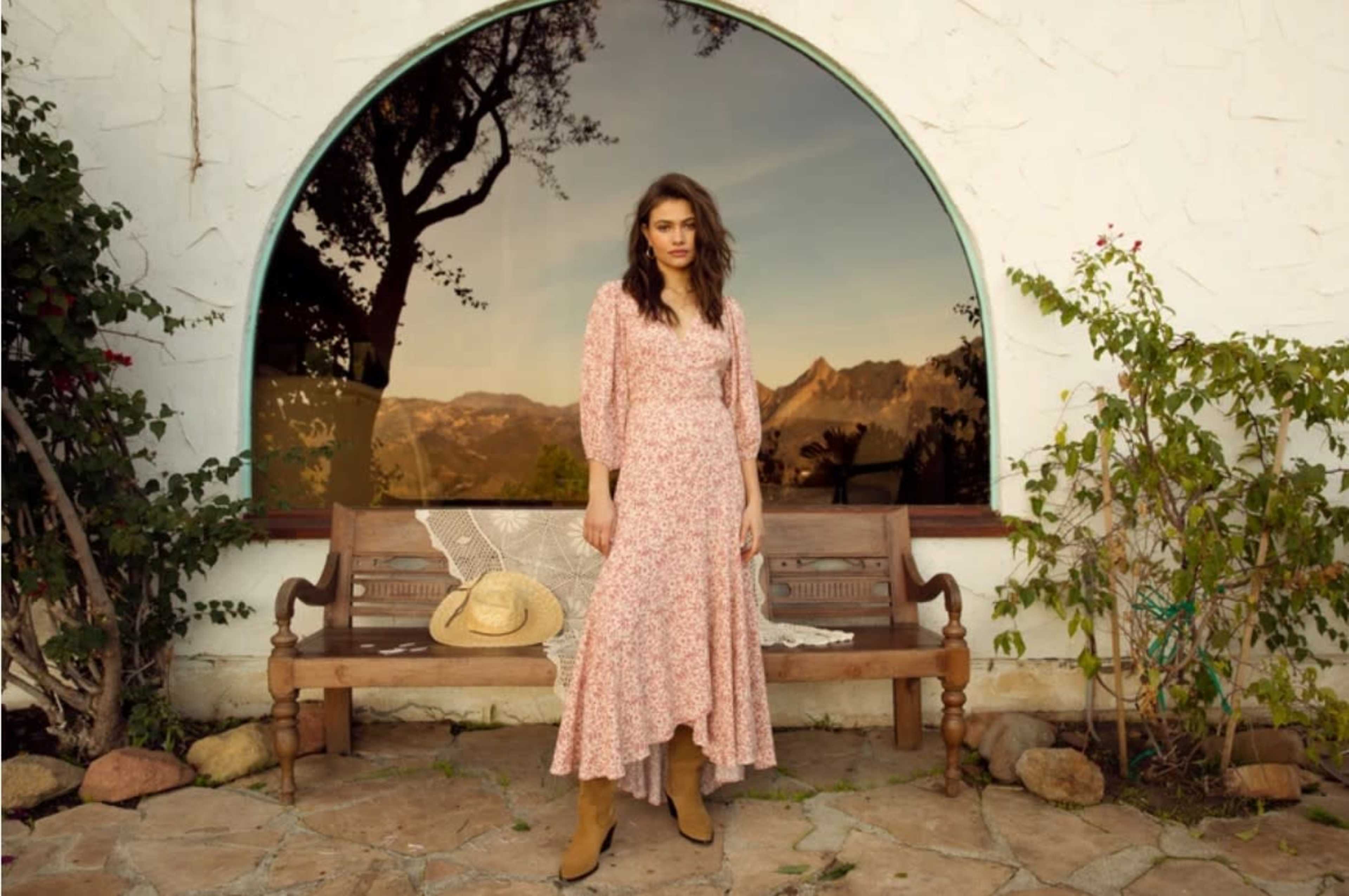 A woman stands on a stone patio in front of a large arched window, wearing a floral dress and brown boots, with a straw hat nearby.