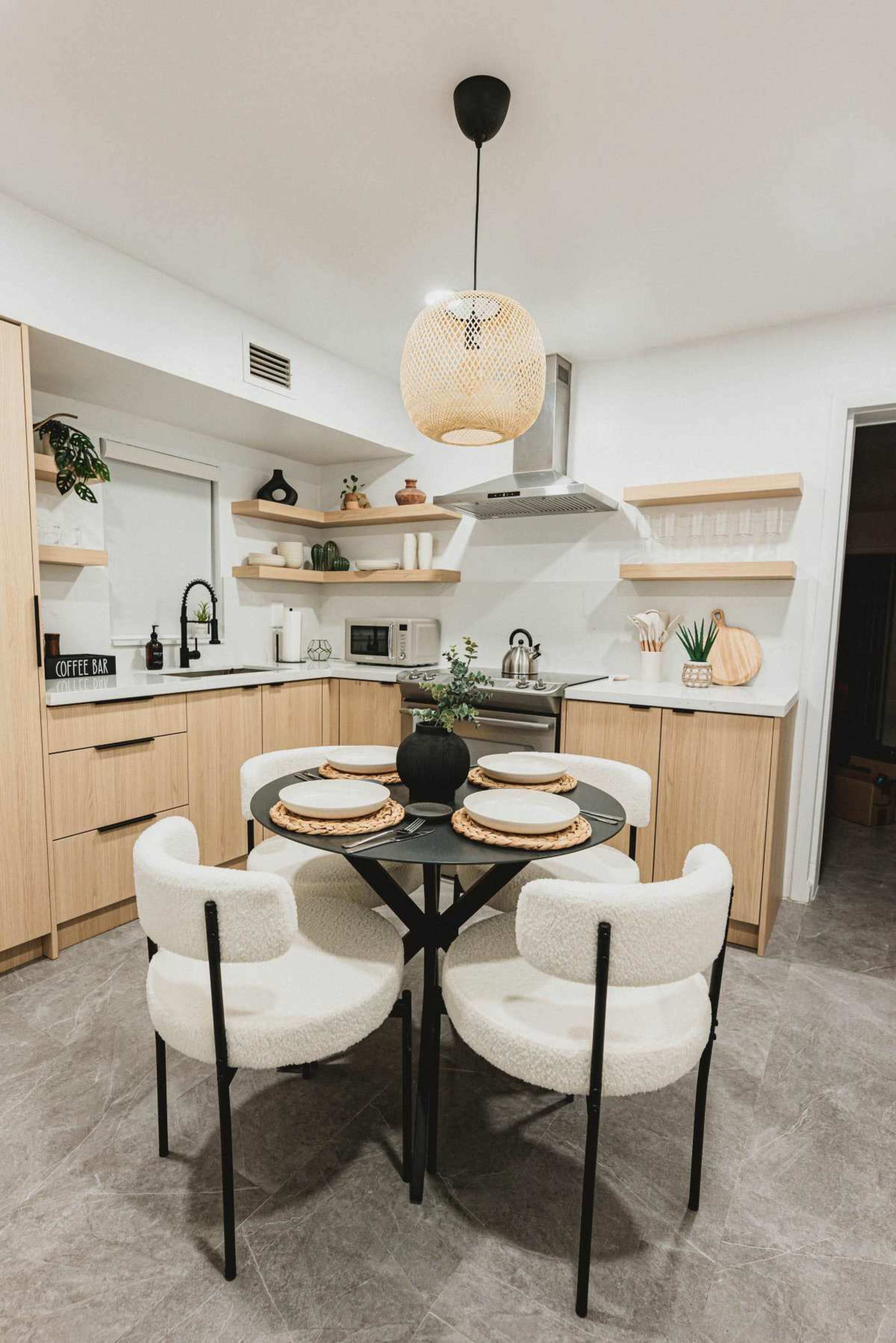 The image shows a modern kitchen with light wood cabinetry, a round black dining table set for four, and a woven pendant light hanging above.