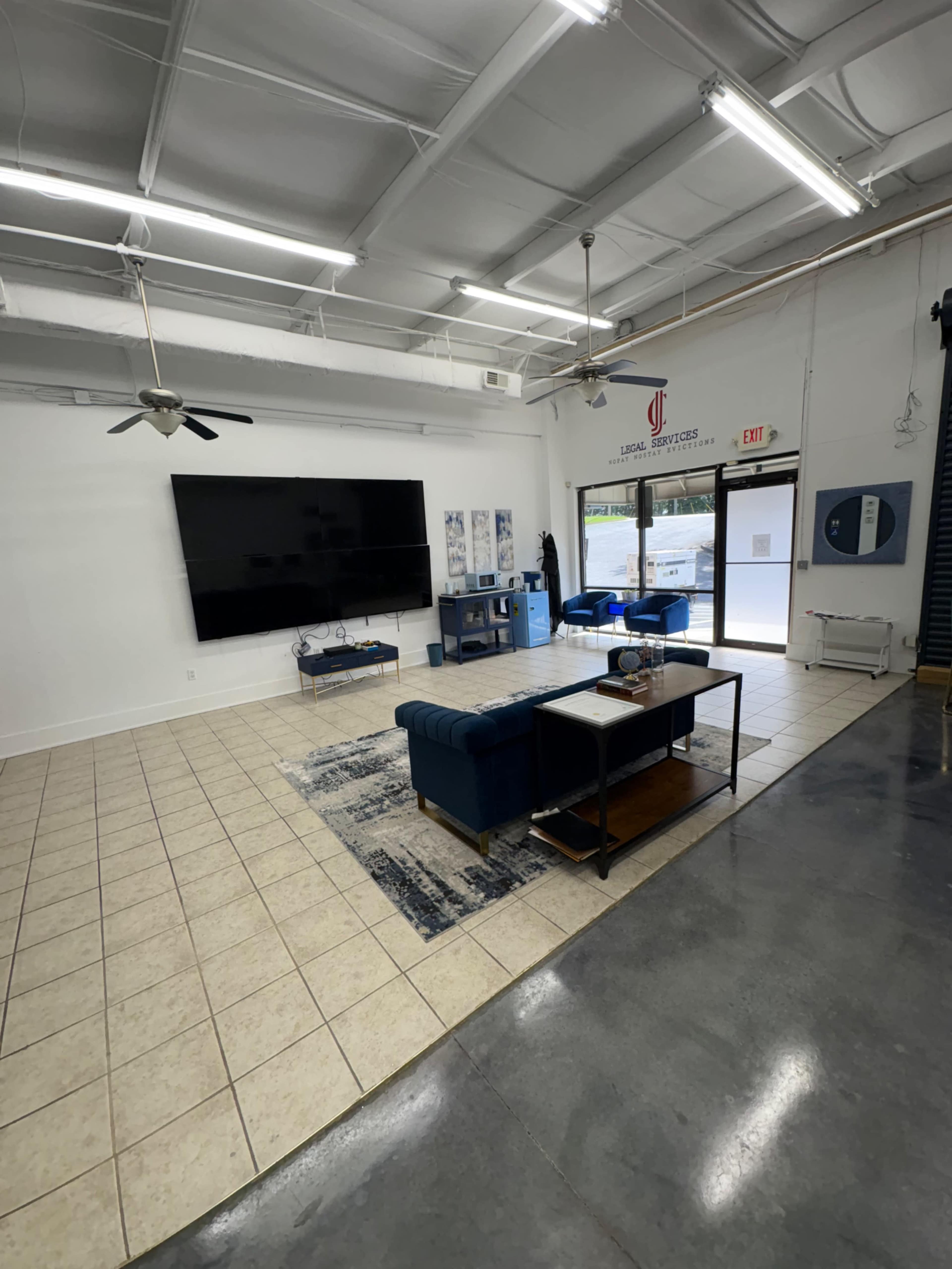The image shows a spacious room with tiled flooring, featuring a large wall-mounted television, a coffee table, and blue seating arrangements, along with commercial windows visible in the background.