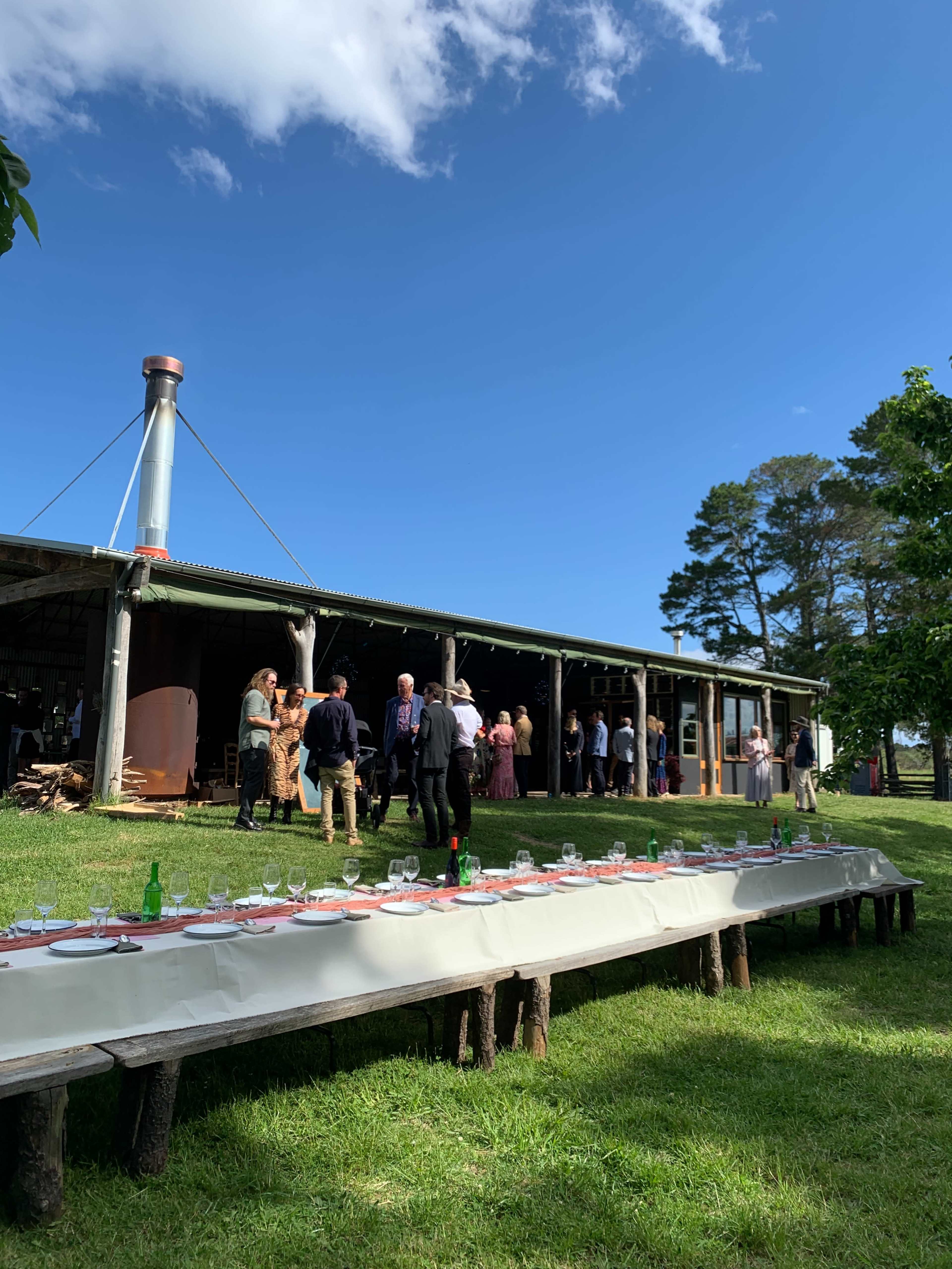 A long table is set outside in front of a rustic building, while guests mingle nearby under a clear blue sky.