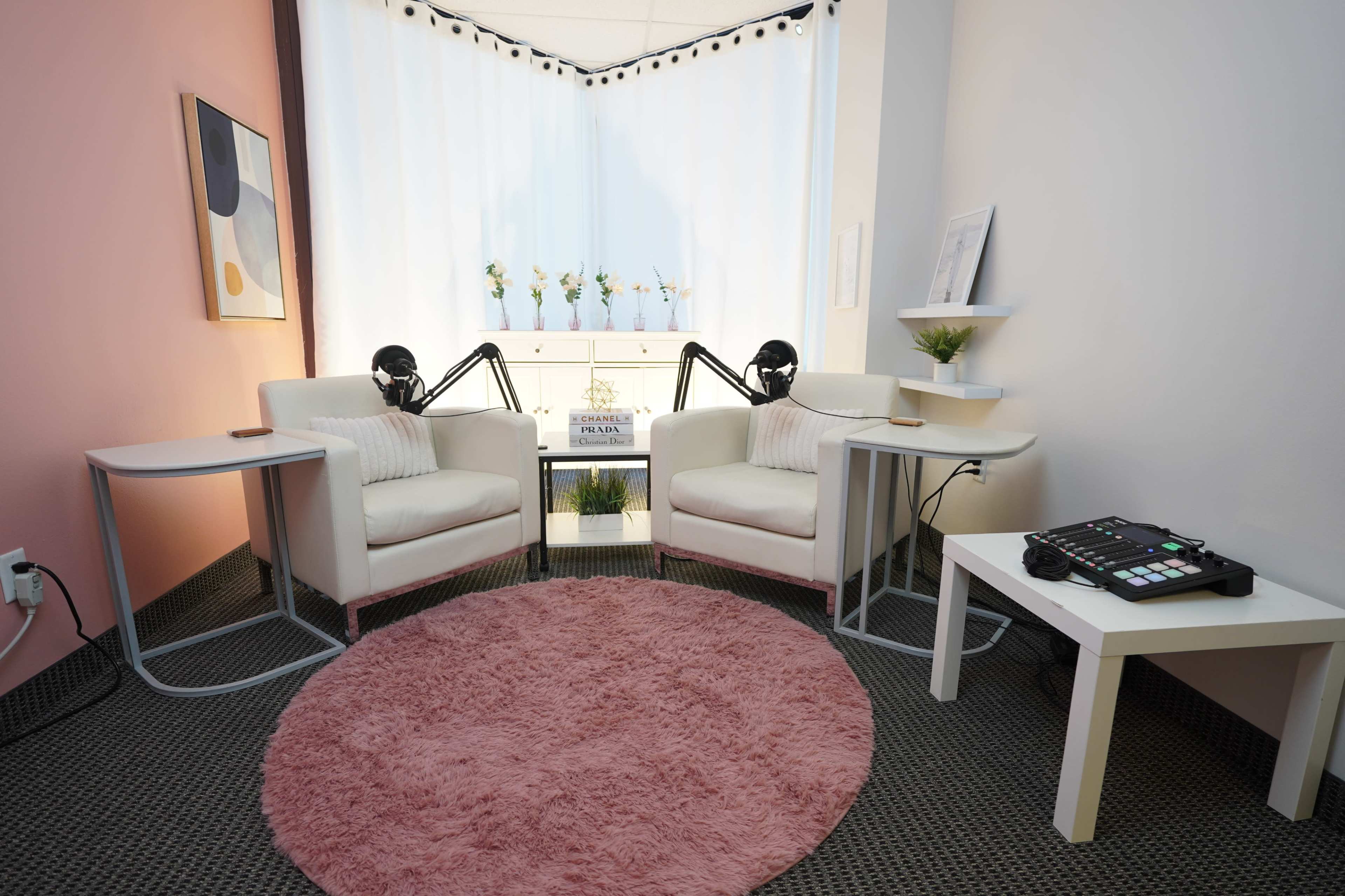 The image shows a small, inviting recording space with two white chairs, a pink rug, and audio equipment on a side table.