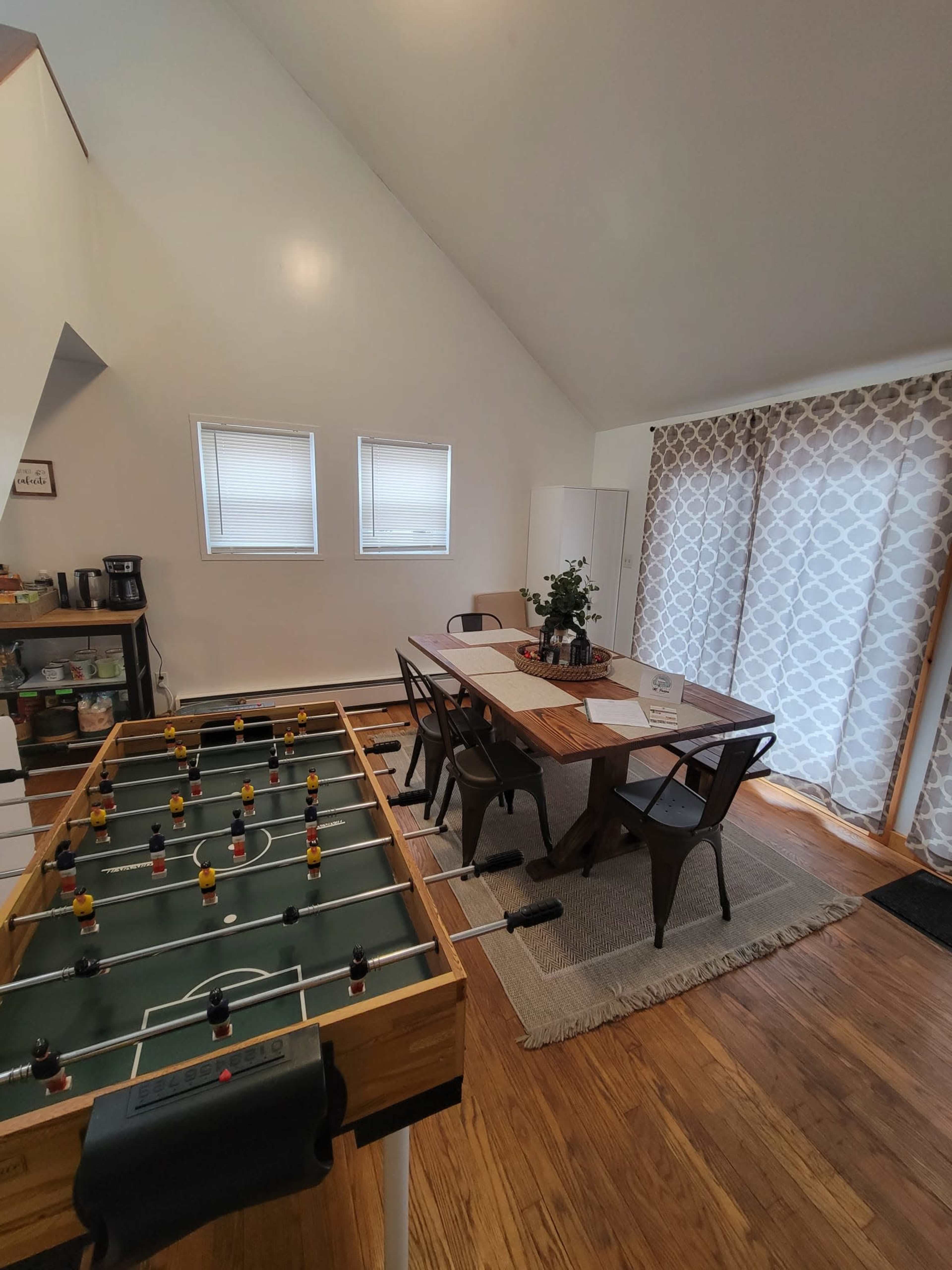 A foosball table is positioned beside a dining area with a wooden table and chairs, where large windows allow natural light to enter.