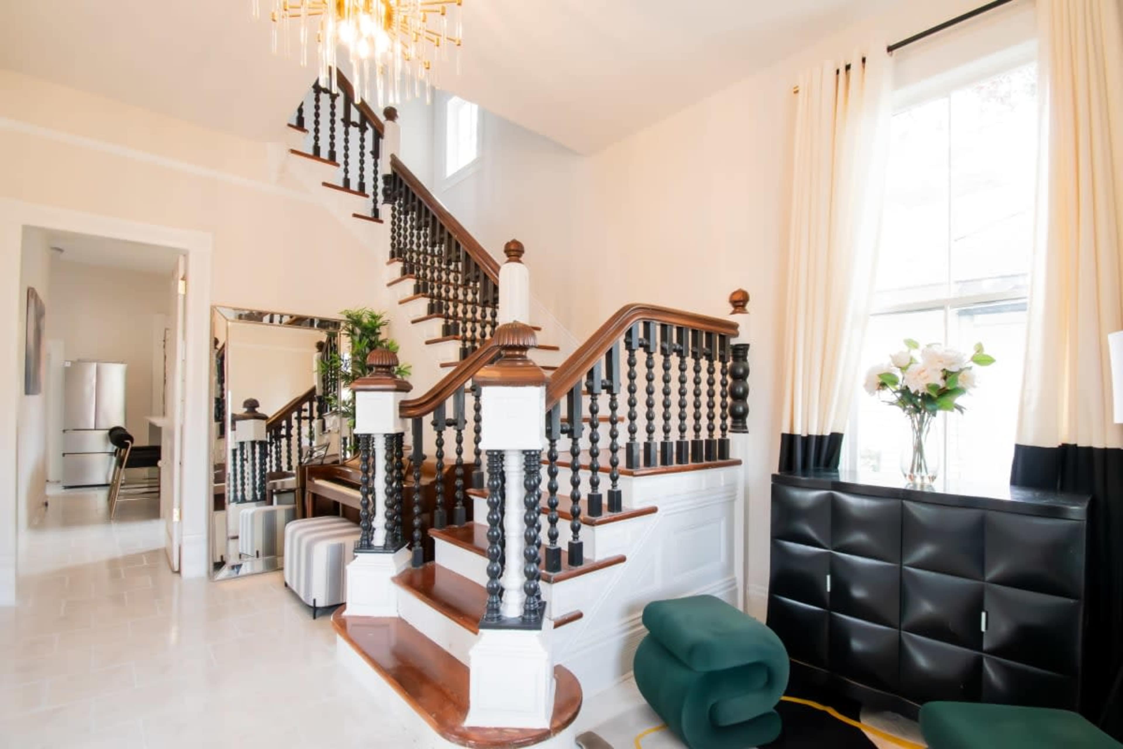 A well-lit foyer featuring a wooden staircase with ornate banisters, a seating area, and large windows with long curtains.