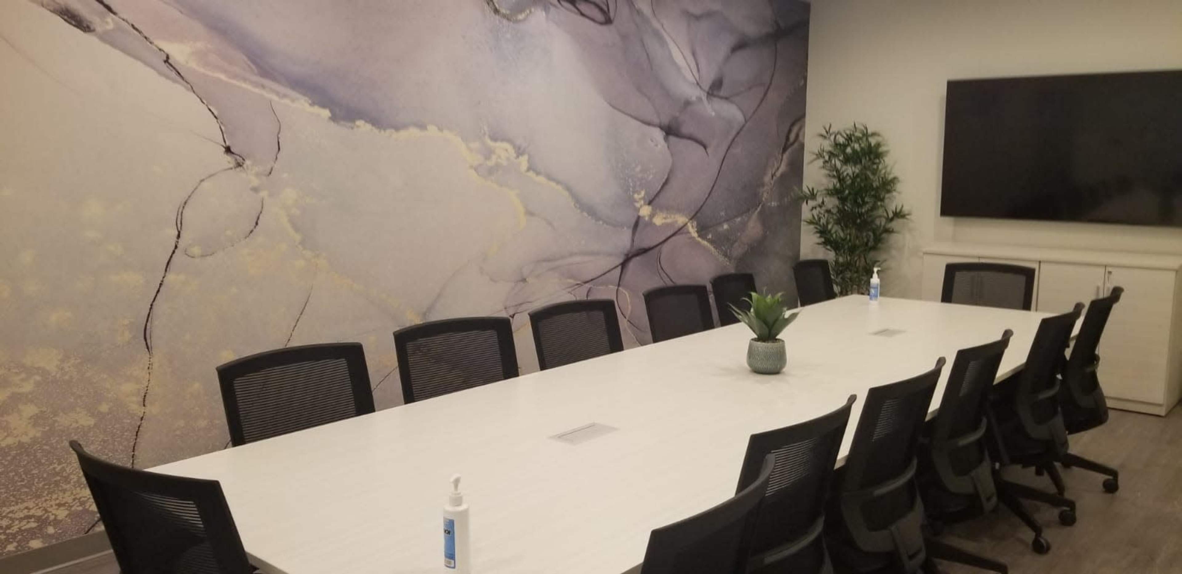 A modern conference room features a long white table surrounded by black chairs, with a decorative plant and a wall adorned with an abstract design.