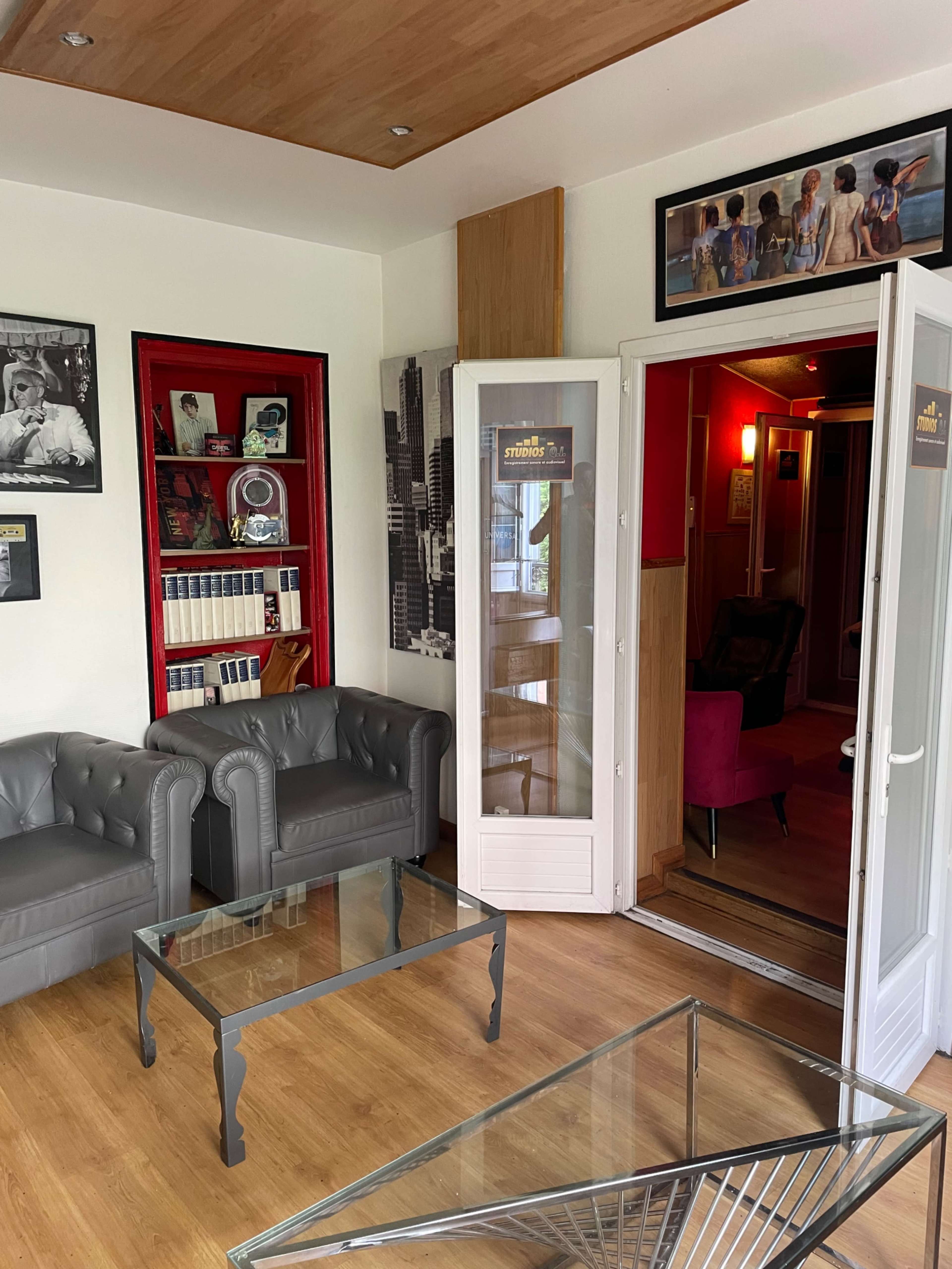 A stylish lounge area featuring two gray tufted sofas, a glass coffee table, and a bookshelf, with an open door leading to a hallway.