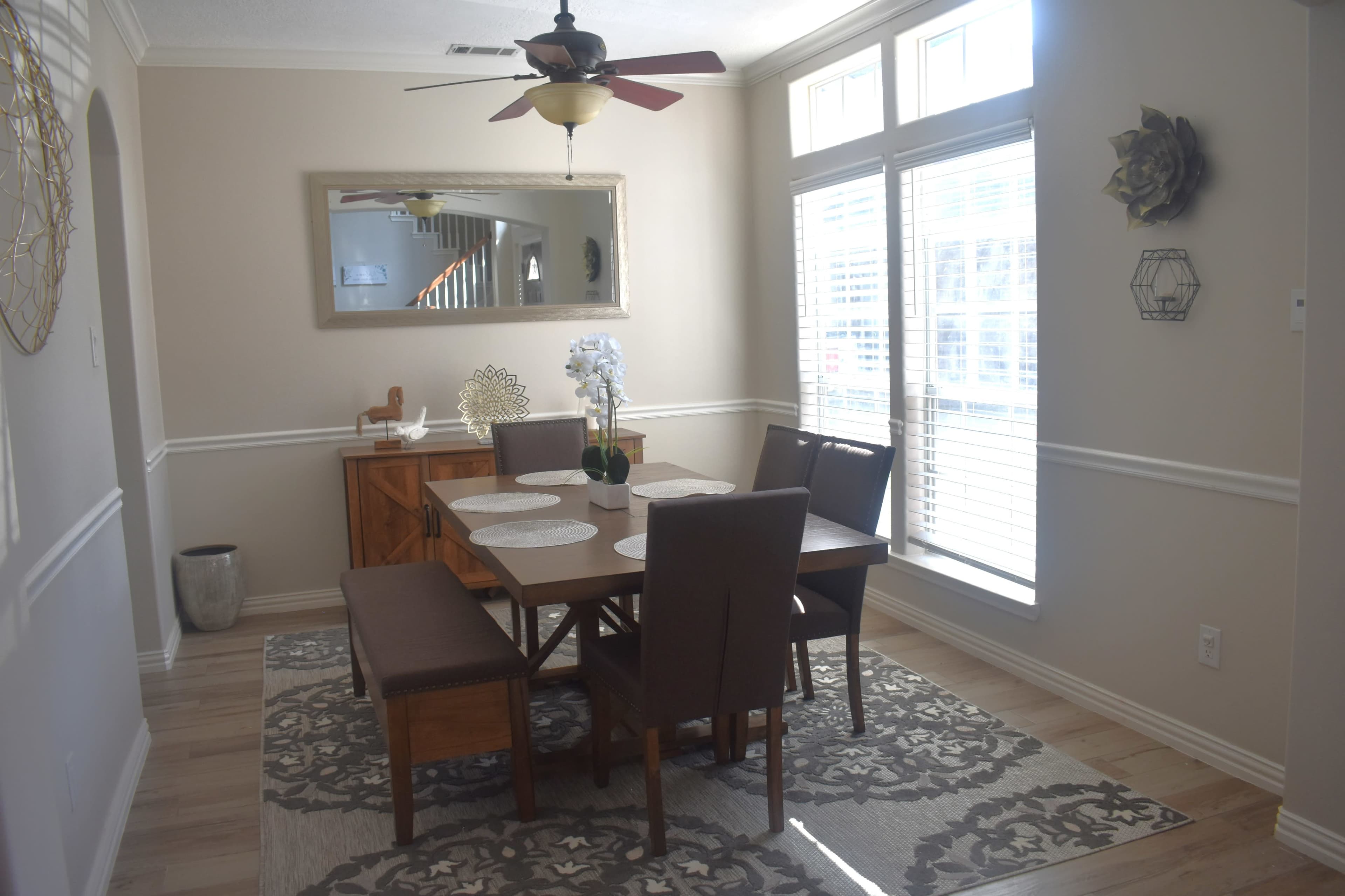 A dining area features a wooden table with upholstered chairs, a decorative mirror, and sunlight streaming through large windows.