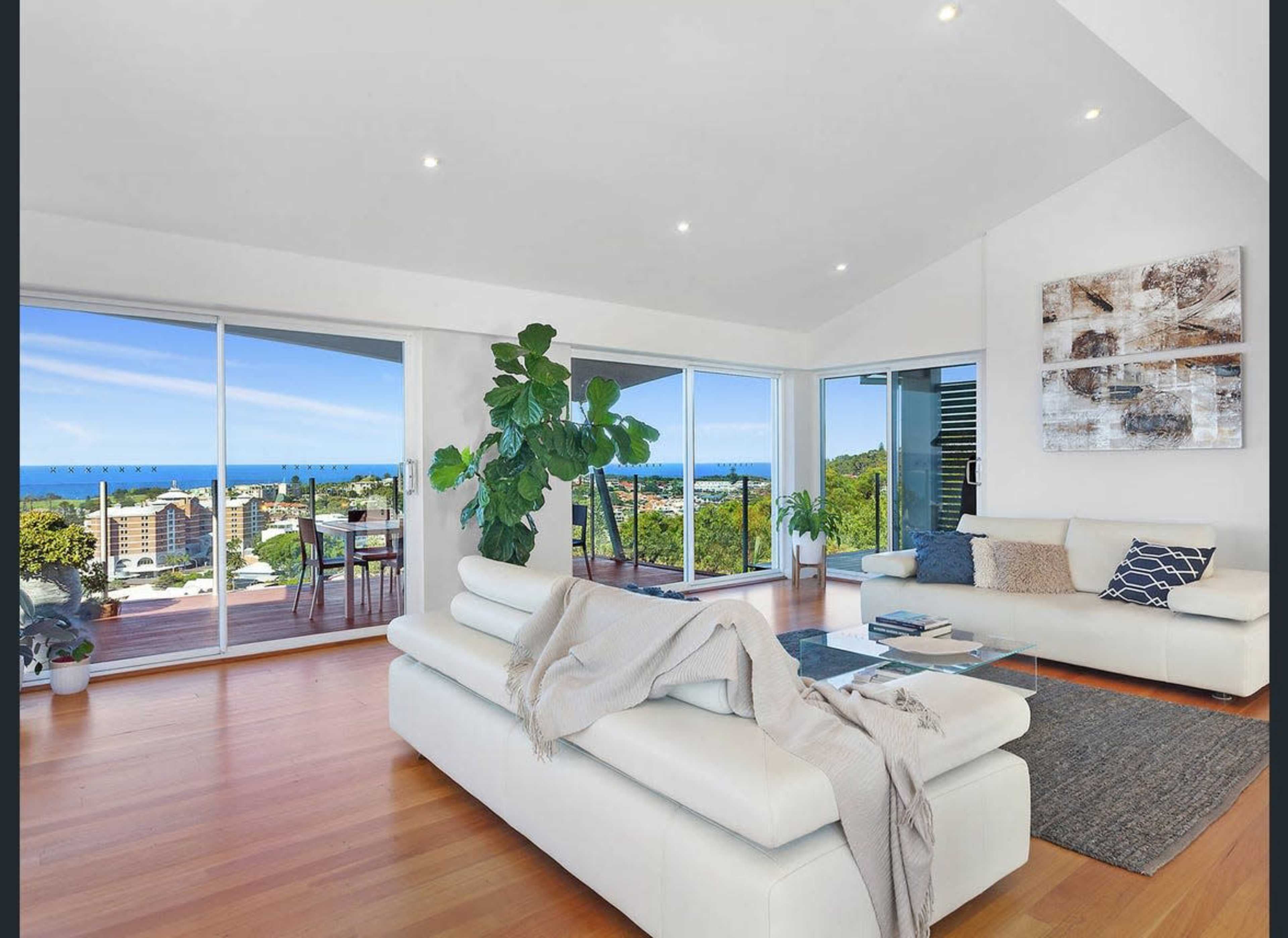 The image shows a modern living room with large windows offering a view of the ocean and a balcony area, featuring a white sofa, a large plant, and wooden flooring.