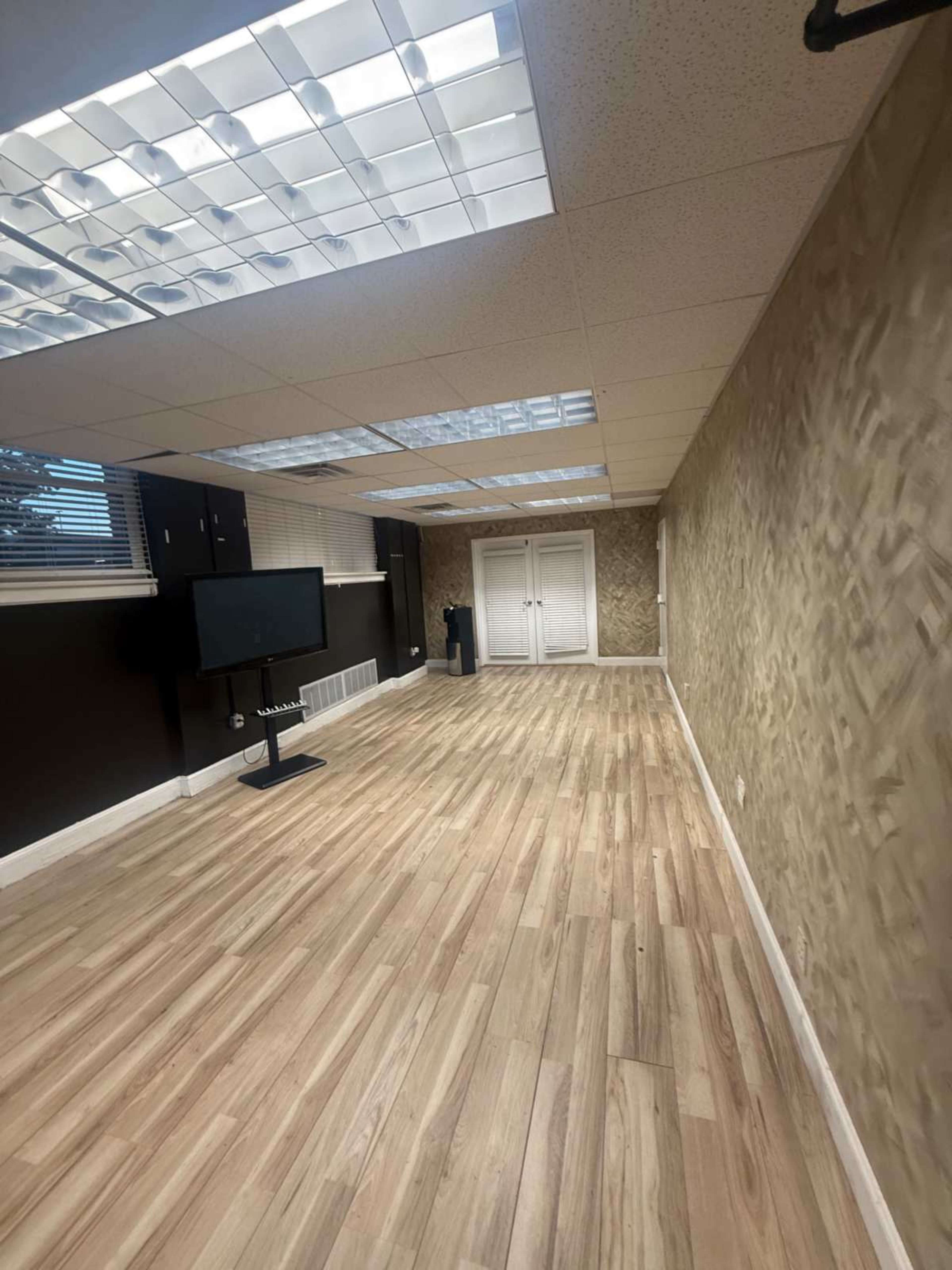 The image shows a spacious, empty room with wooden flooring, light-colored walls, and a television mounted on a stand against one wall.