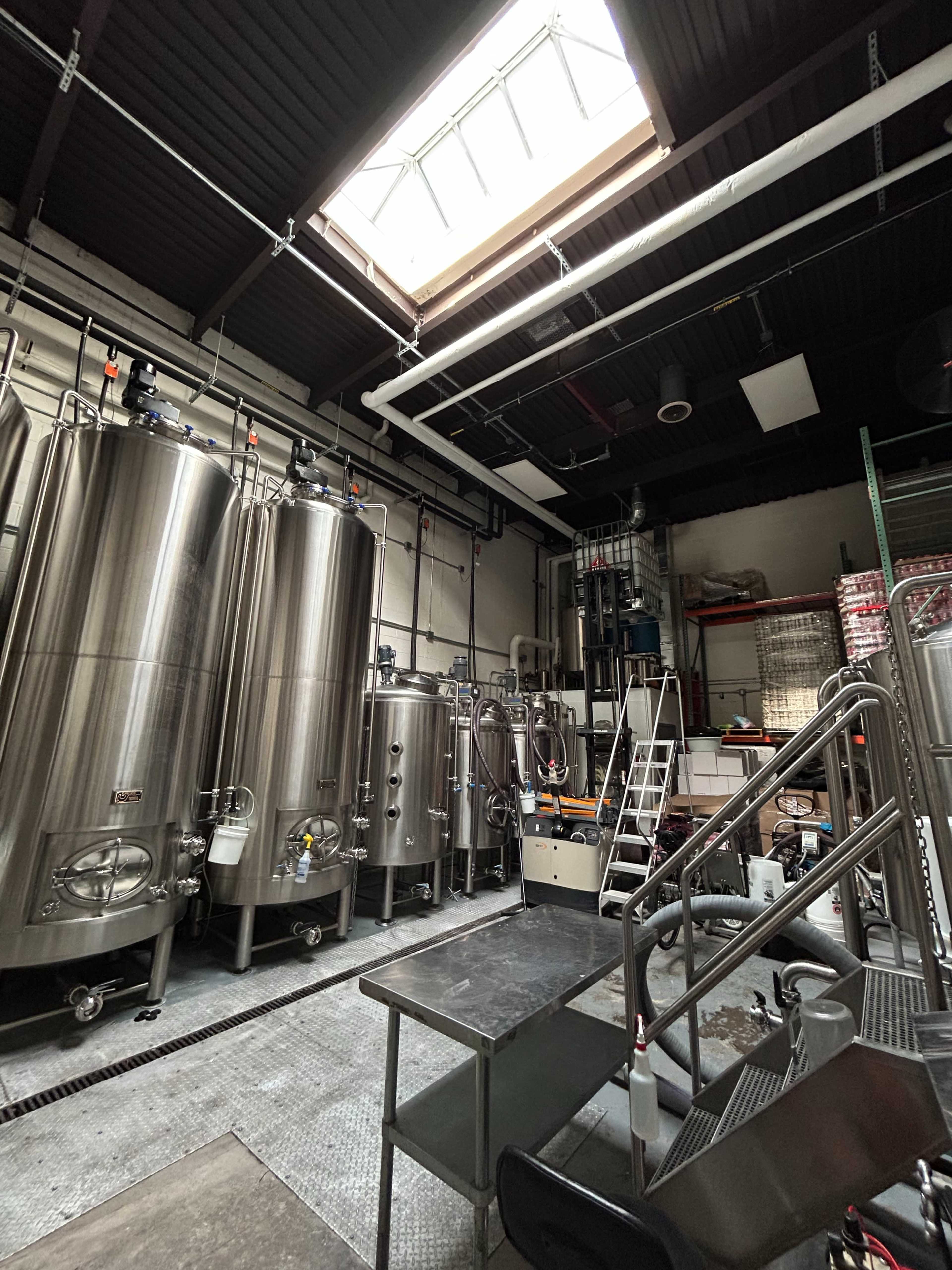 The image shows a brew room with large stainless steel fermentation tanks and industrial equipment, featuring a skylight above.
