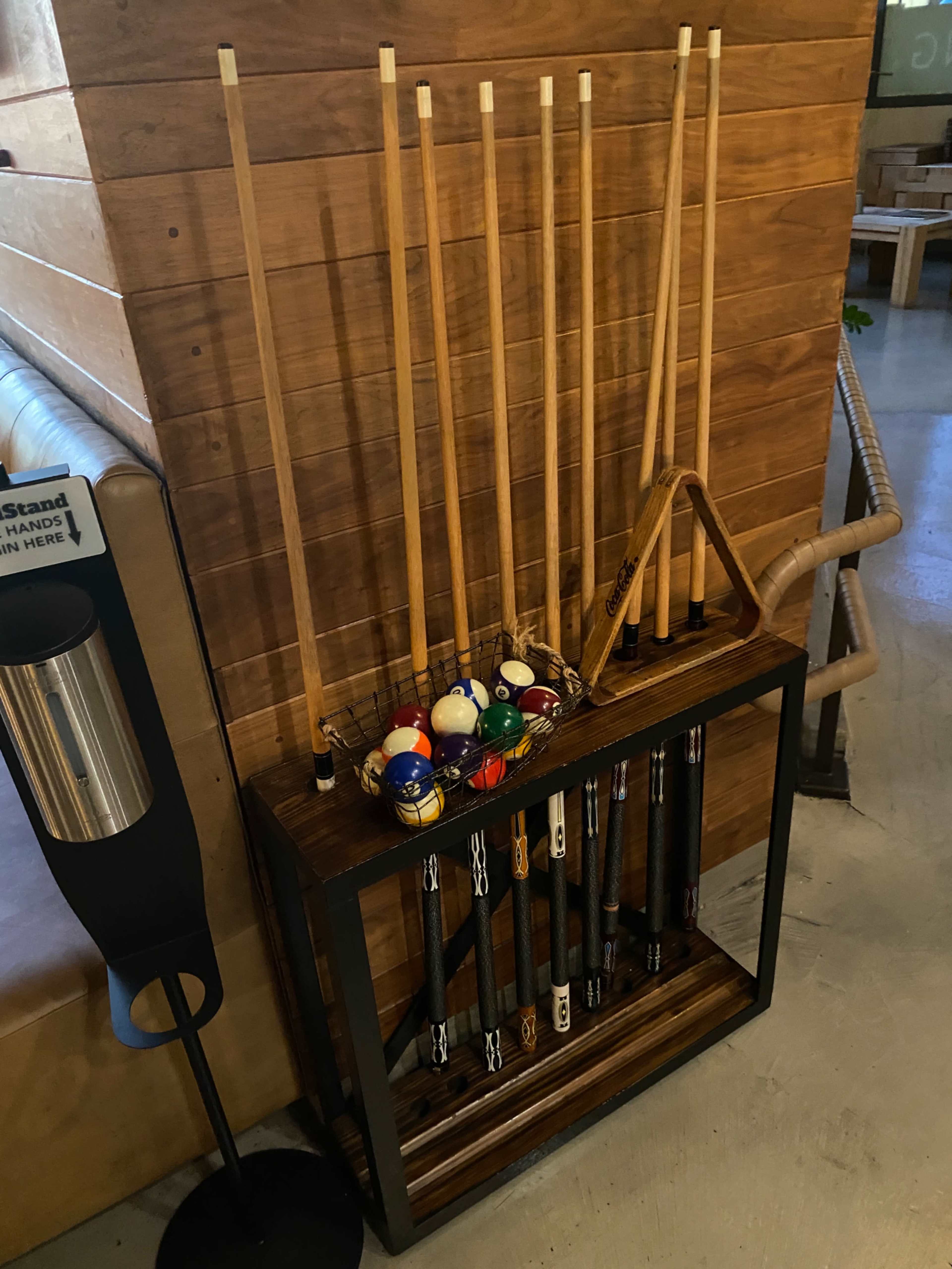 A rack holds several pool cues and a triangle rack above a wooden shelf containing various colored pool balls.