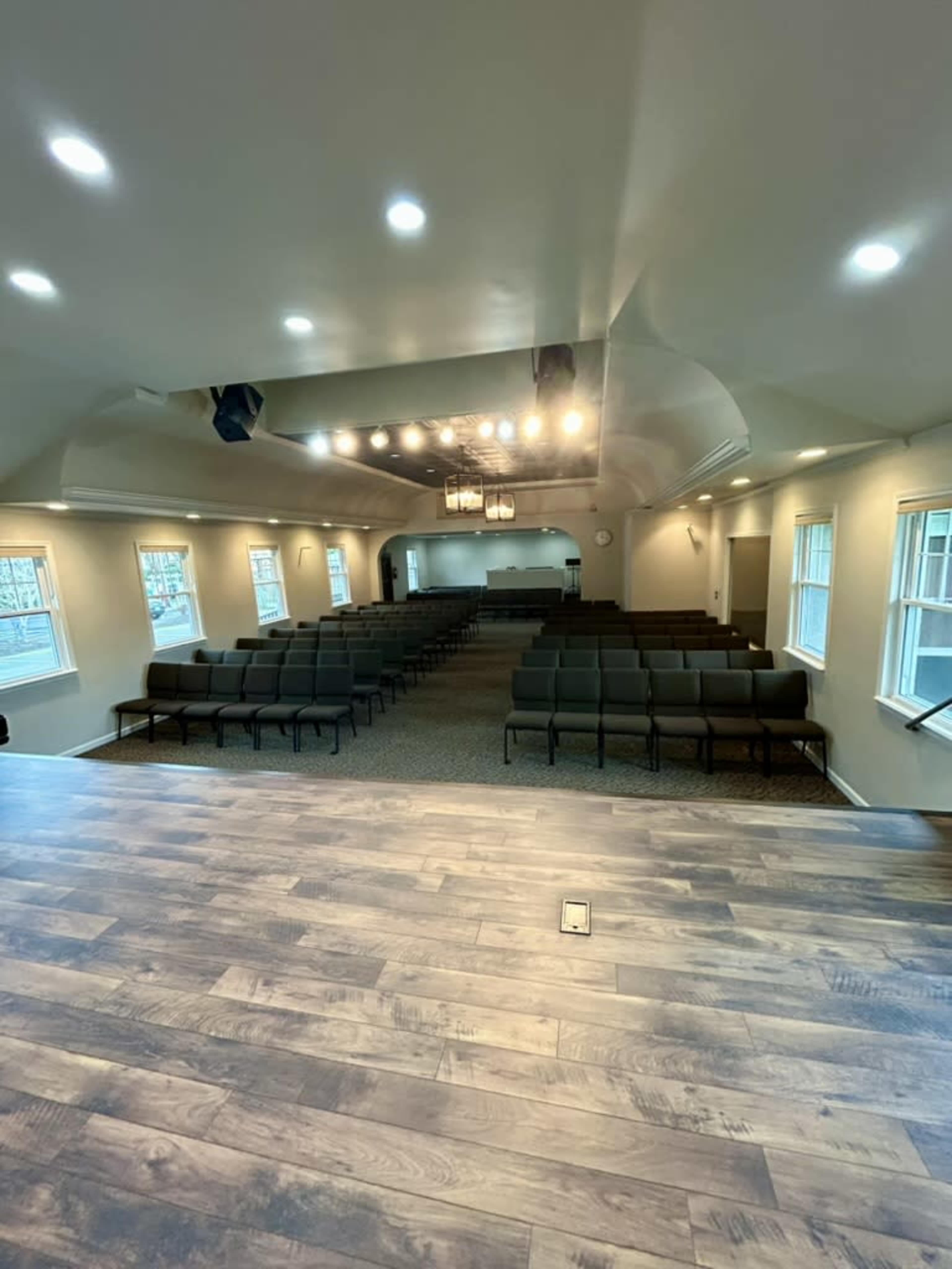 The image shows an indoor space with rows of empty seating facing a stage, illuminated by overhead lights.