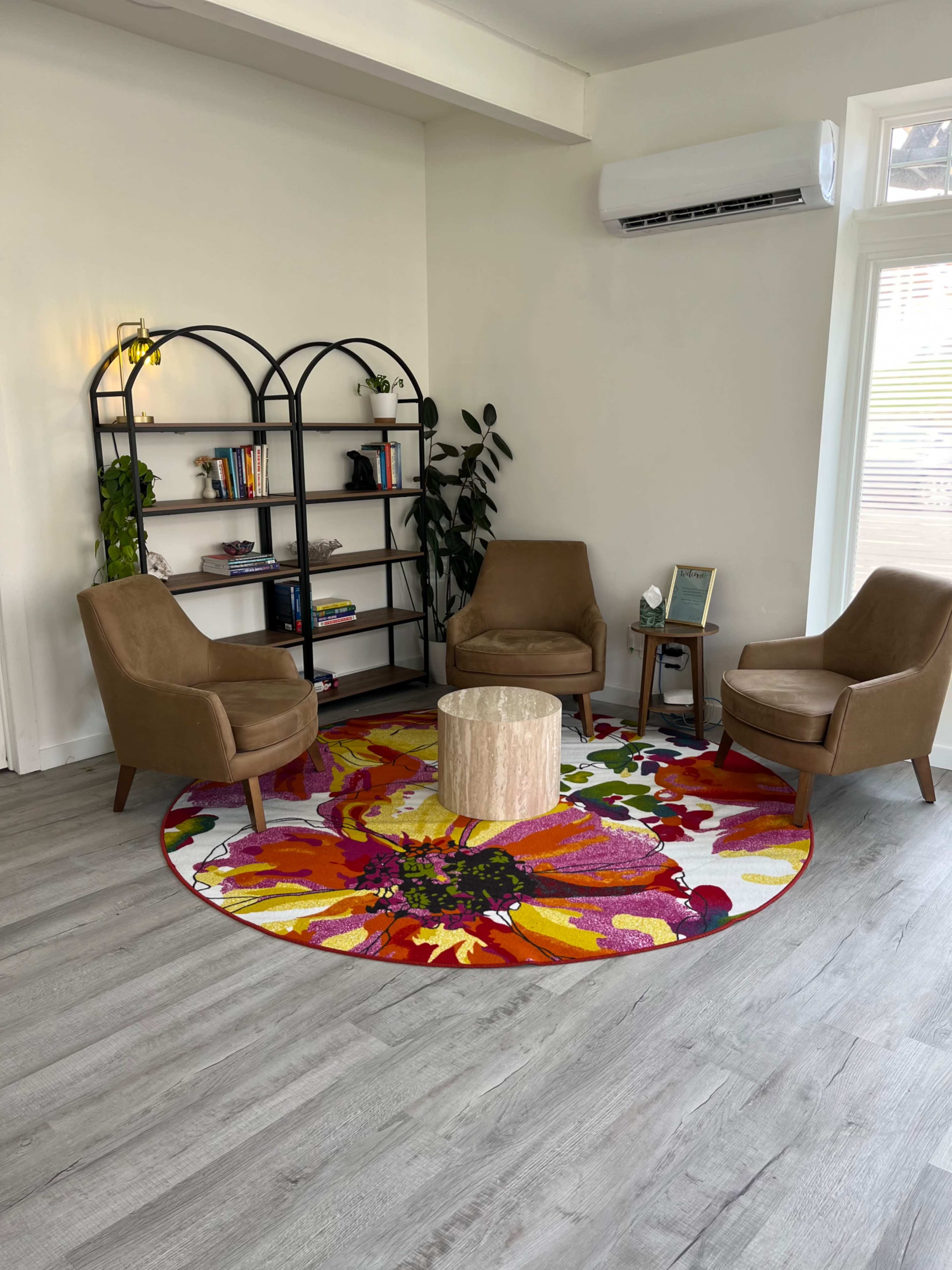 The image shows a brightly decorated seating area with two brown chairs, a wooden coffee table, and a circular, colorful floral rug, surrounded by a bookshelf and a potted plant.