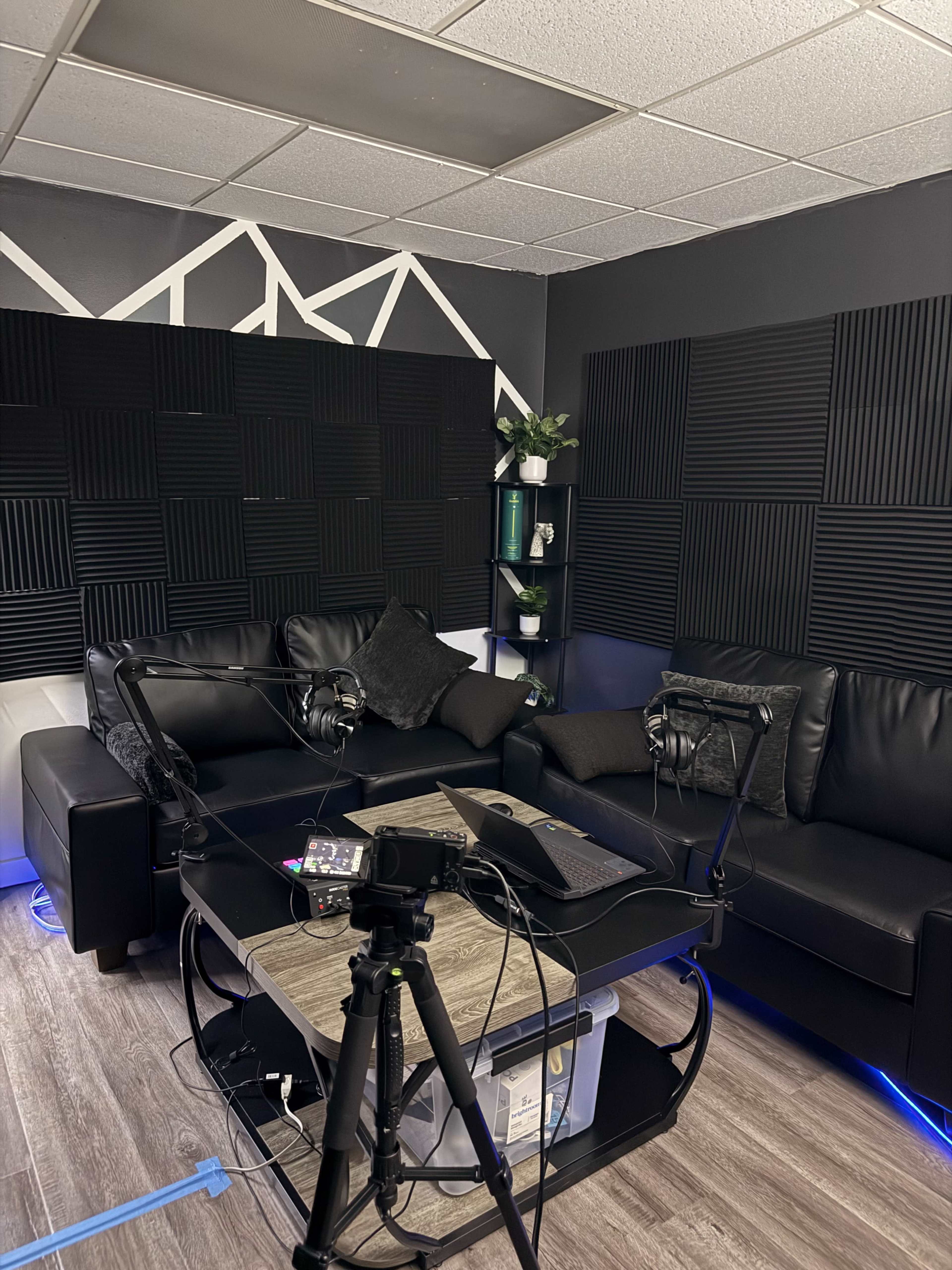 The image shows a small recording studio with black soundproof panels on the walls, a black leather couch, and a coffee table equipped with microphones and recording equipment.