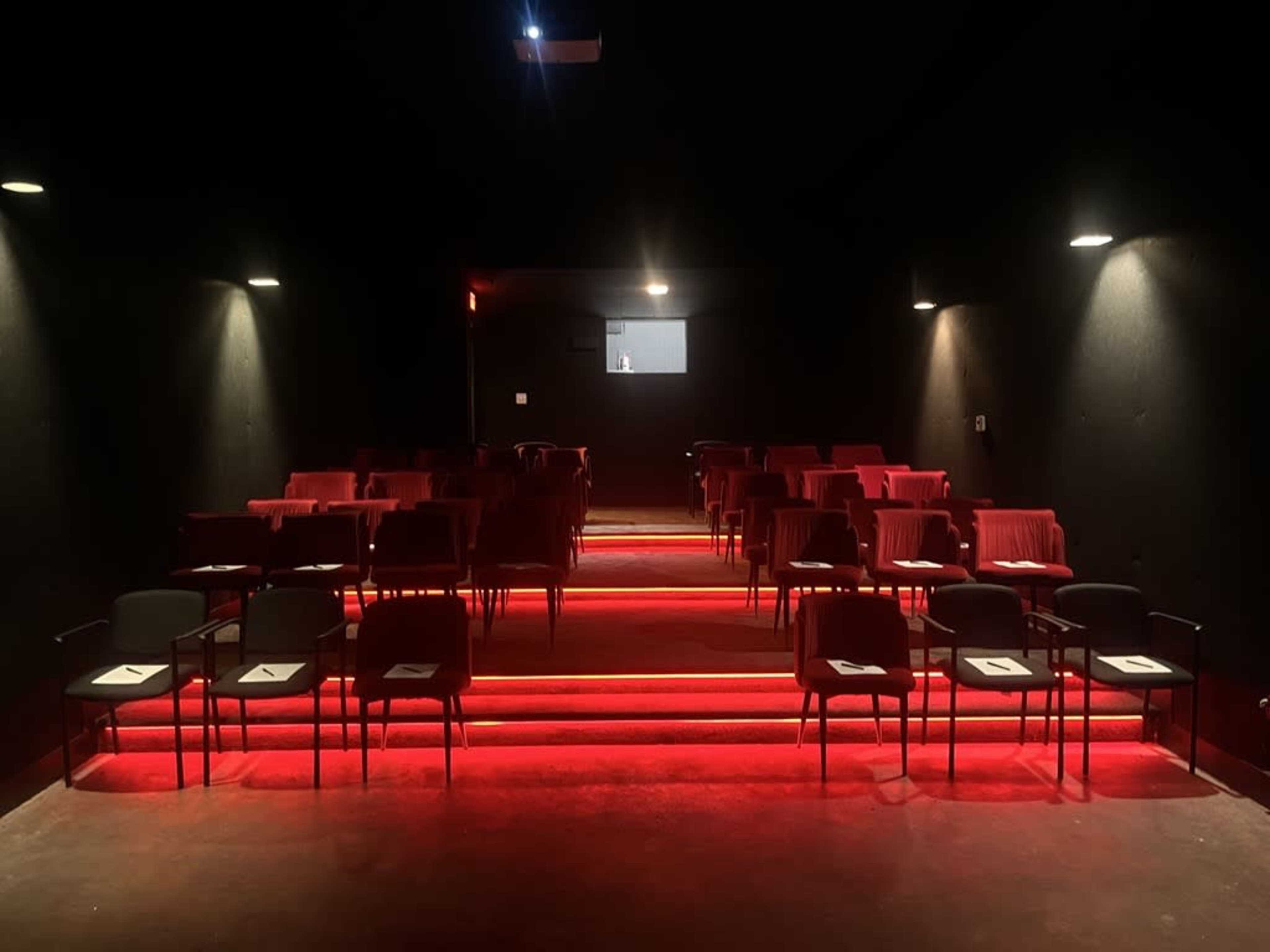 A dimly lit theater features red-highlighted seating arranged on tiered steps.