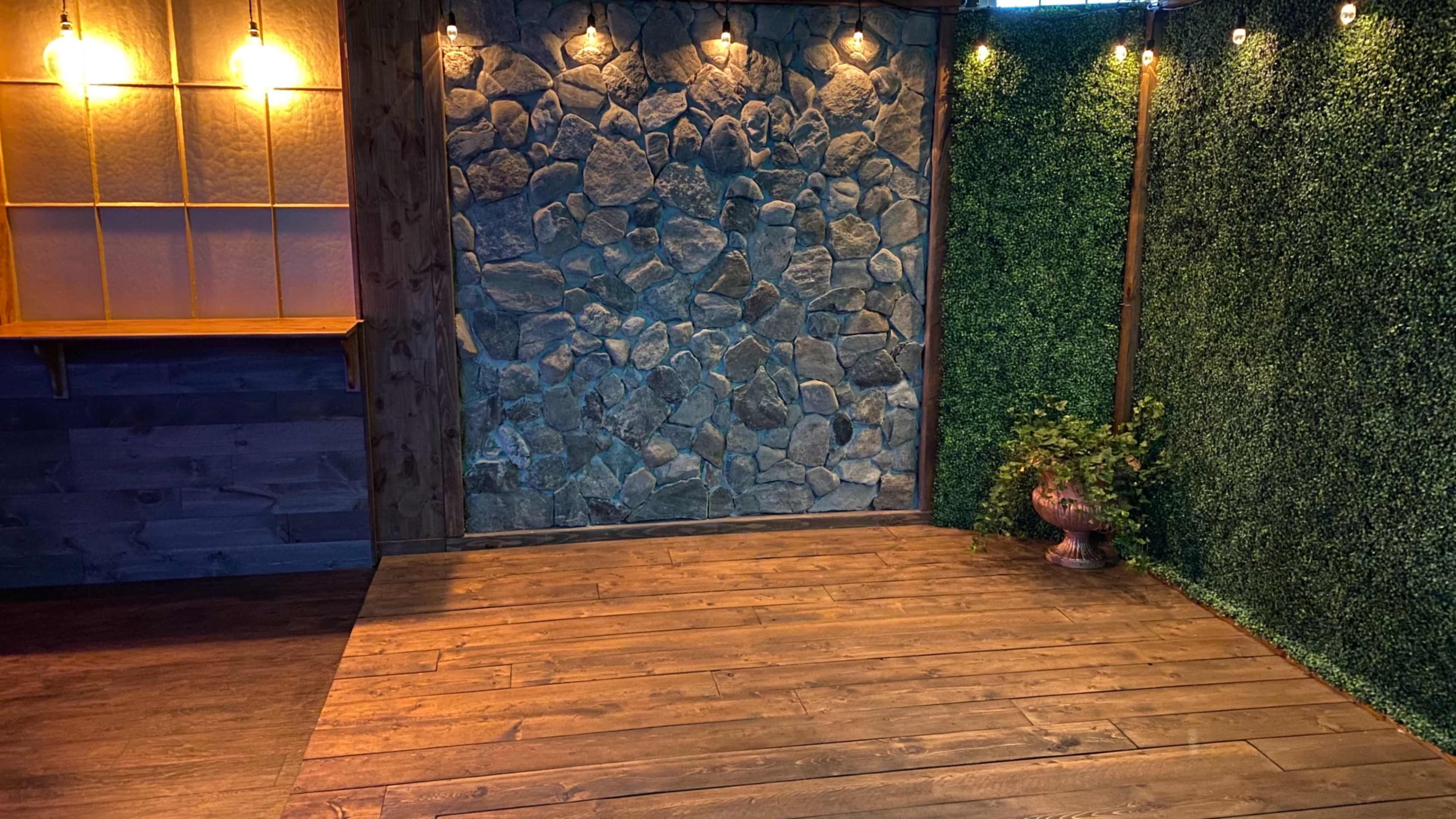 A wooden floor area with a stone wall and greenery on one side, illuminated by warm lights.